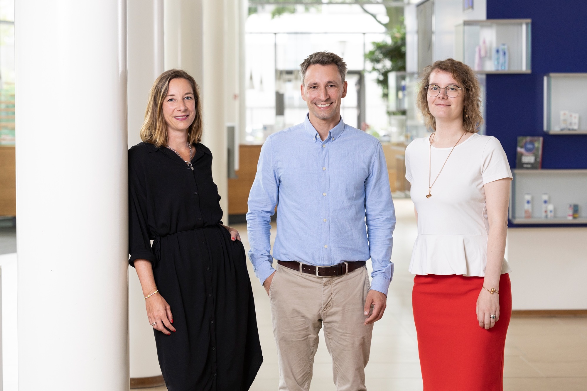 Three people stand indoors in a well-lit, modern space at Hochschule Coburg. A woman in a black dress leans against a white column, while a man in a blue shirt and beige pants stands in the center. Another woman, wearing a white top and red skirt, is positioned on the right.