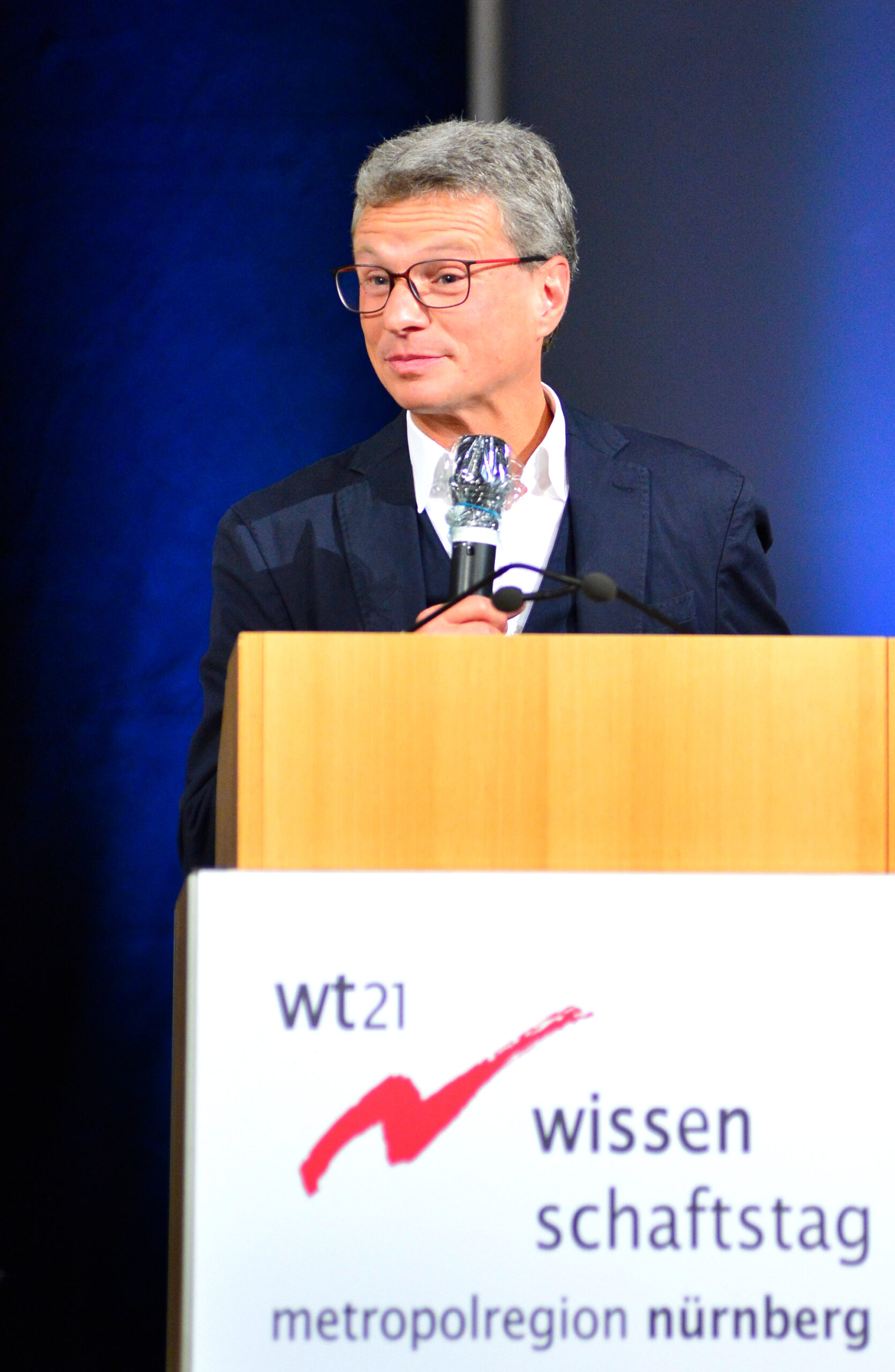 Eine Person mit Brille spricht in ein Mikrofon an einem Podium, das mit dem Schild „Wissenschaftstag Metropolregion Nürnberg“ mit einem roten Häkchen-Logo geschmückt ist. Der dunkelblaue und graue Hintergrund ergänzt diese Veranstaltung. Er spiegelt den kollaborativen Geist wider, der bei akademischen Zusammenkünften der Hochschule Coburg herrscht.