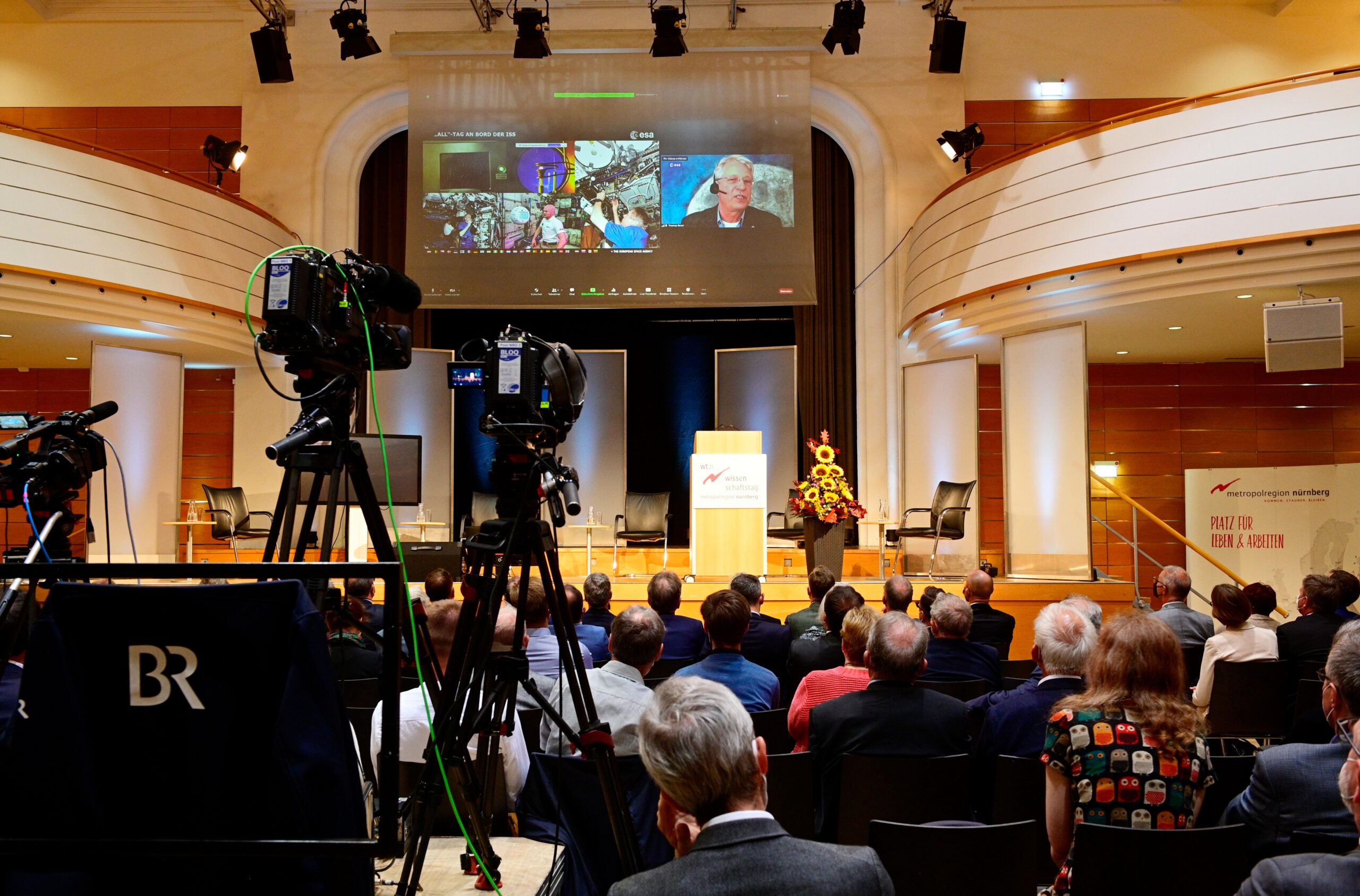 Das Publikum sitzt in einem modernen Hörsaal der Hochschule Coburg, während Kameras eine Veranstaltung filmen. Auf einem großen Bildschirm sind mehrere Personen in einem Videoanruf zu sehen. Die Bühne ist mit einem Blumenarrangement und einem Podium geschmückt und schafft so eine Atmosphäre der Innovation und Vernetzung.