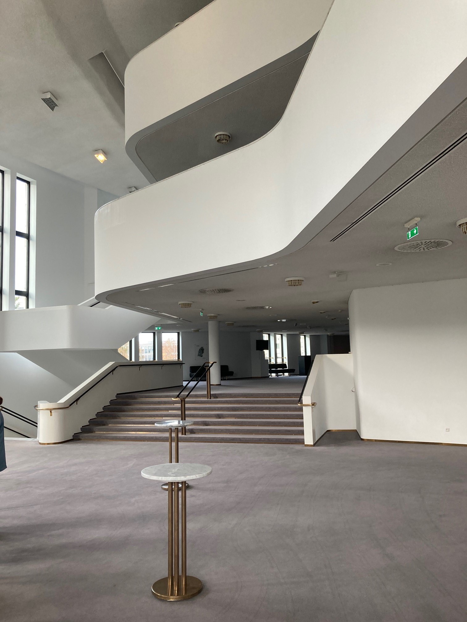 Großzügiges, modernes Interieur der Hochschule Coburg mit geschwungener weißer Treppe, geschwungenem Balkon und minimalistischem Design. Hohe Fenster lassen Tageslicht herein und im Vordergrund stehen verstreut kleine runde Tische mit goldenen Untergestellen.