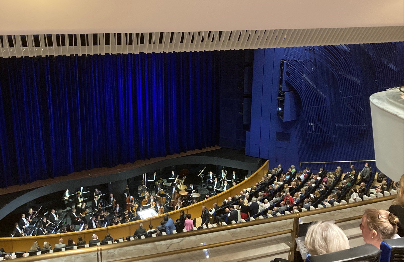 Das Publikum sitzt in einem modernen Hörsaal der Hochschule Coburg, an dessen Vorderseite sich ein Orchestergraben befindet. Die Musiker bereiten sich auf ihr Spiel vor, während die Zuschauer zuschauen. Auf der Bühne sind teilweise blaue Vorhänge zu sehen.