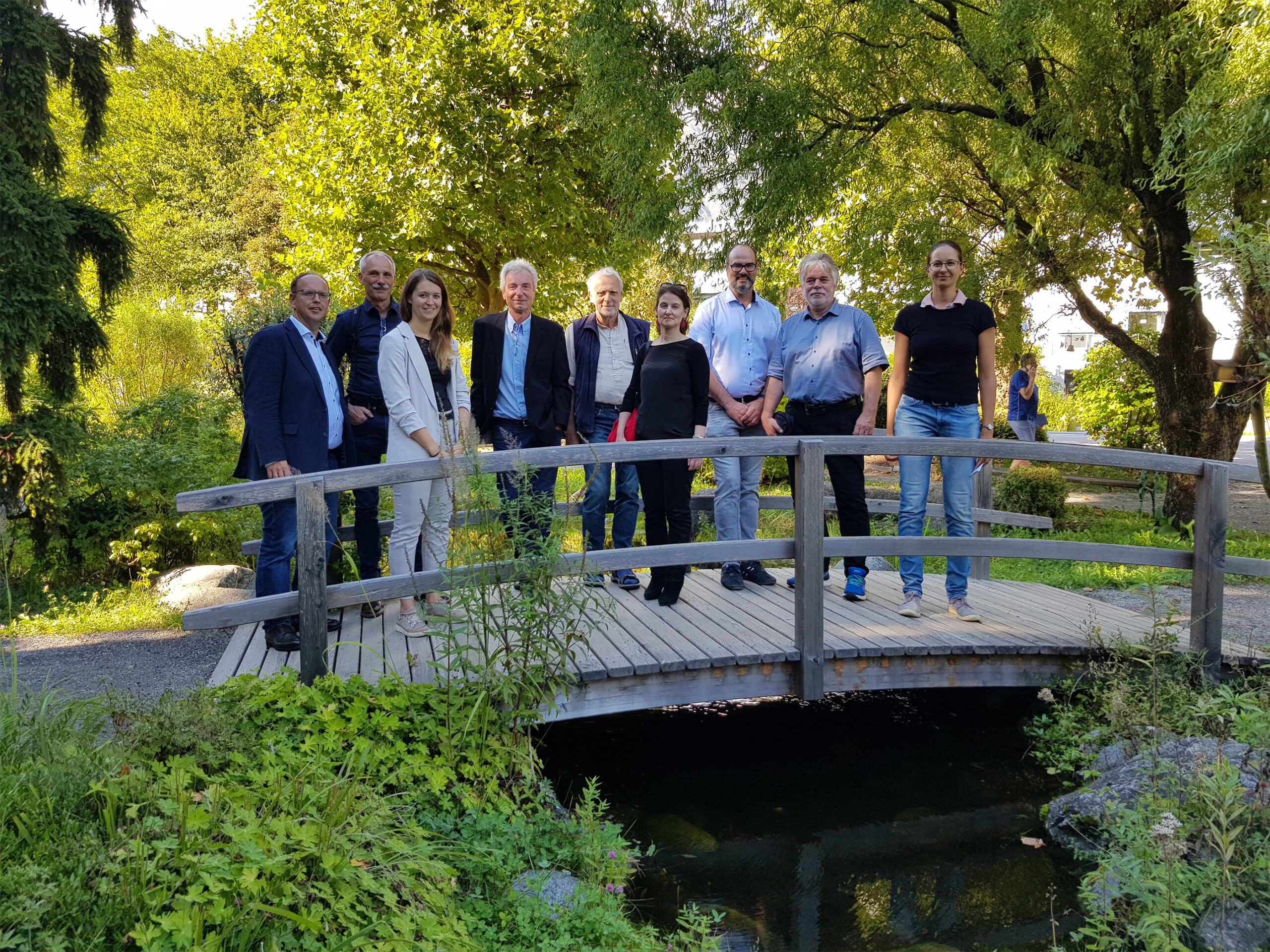 Eine Gruppe von Menschen, möglicherweise Studierende der Hochschule Coburg, steht auf einer kleinen Holzbrücke in einem üppigen Garten. Bäume und Grün umgeben sie, darunter ist ein Teich zu sehen. Lässig gekleidet genießen sie den sonnigen Tag im Freien.