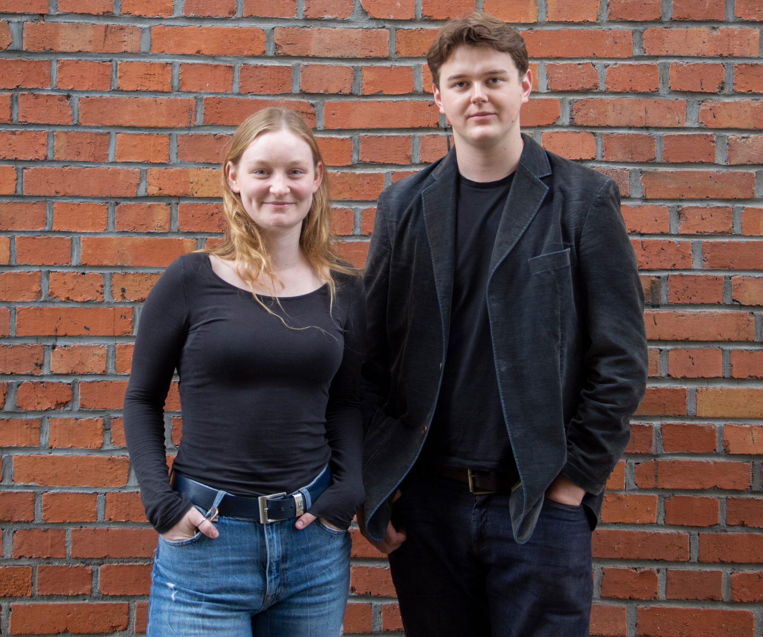 Zwei junge Erwachsene stehen vor einer Backsteinmauer an der Hochschule Coburg. Beide haben helle Haare und tragen legere dunkle Kleidung. Die Person links hat lange Haare, die Person rechts trägt eine Jacke. Beide strahlen ein entspanntes Auftreten aus.