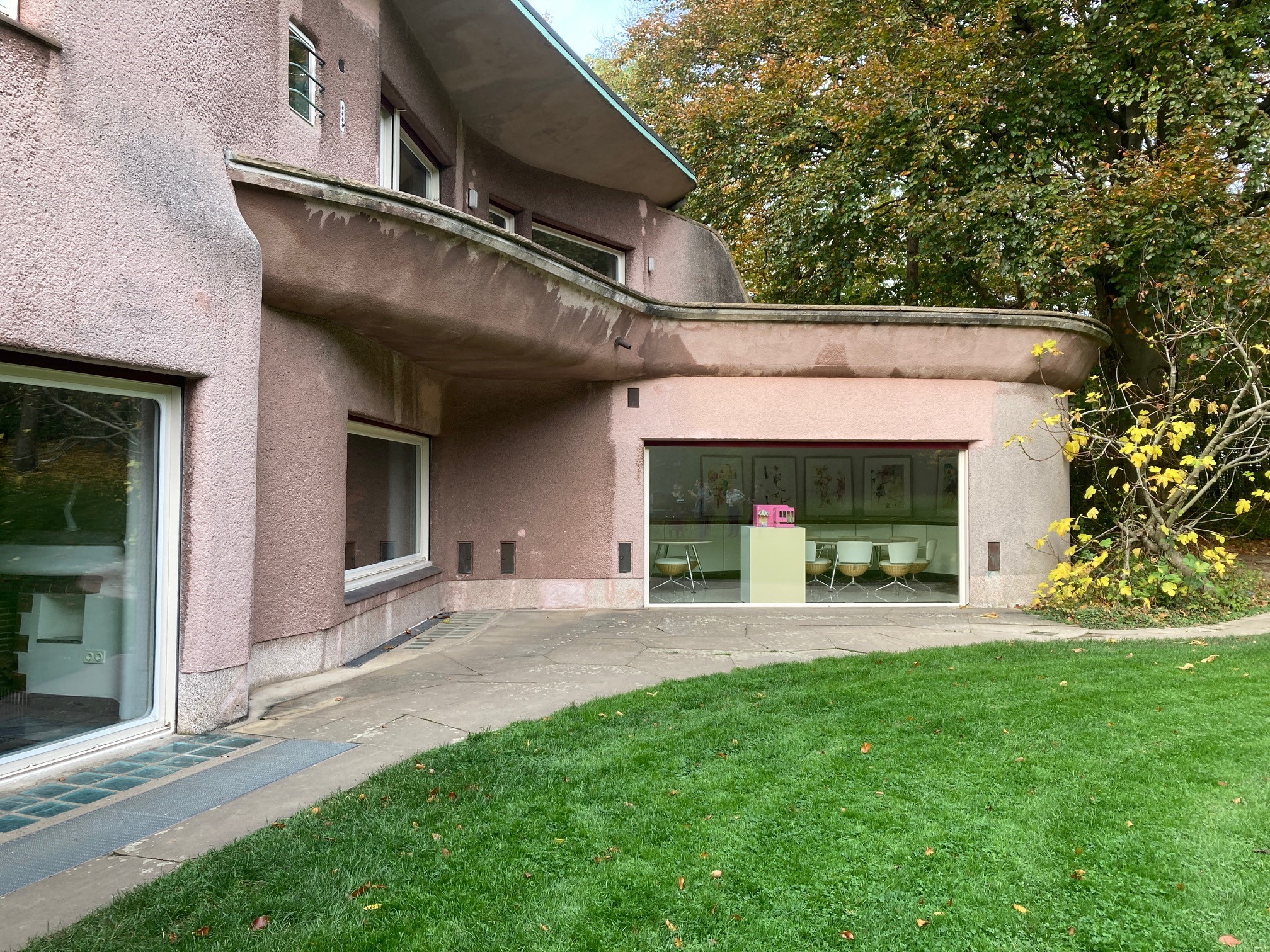 Ein modernes, geschwungenes Gebäude, das an den Architekturstil der Hochschule Coburg erinnert, verfügt über rosafarbene Außenwände, große Fenster und eine Glasschiebetür, die zu einem Raum mit weißen Stühlen führt. Das Gebäude liegt wunderschön vor einem üppigen grünen Rasen mit Laubbäumen im Hintergrund.