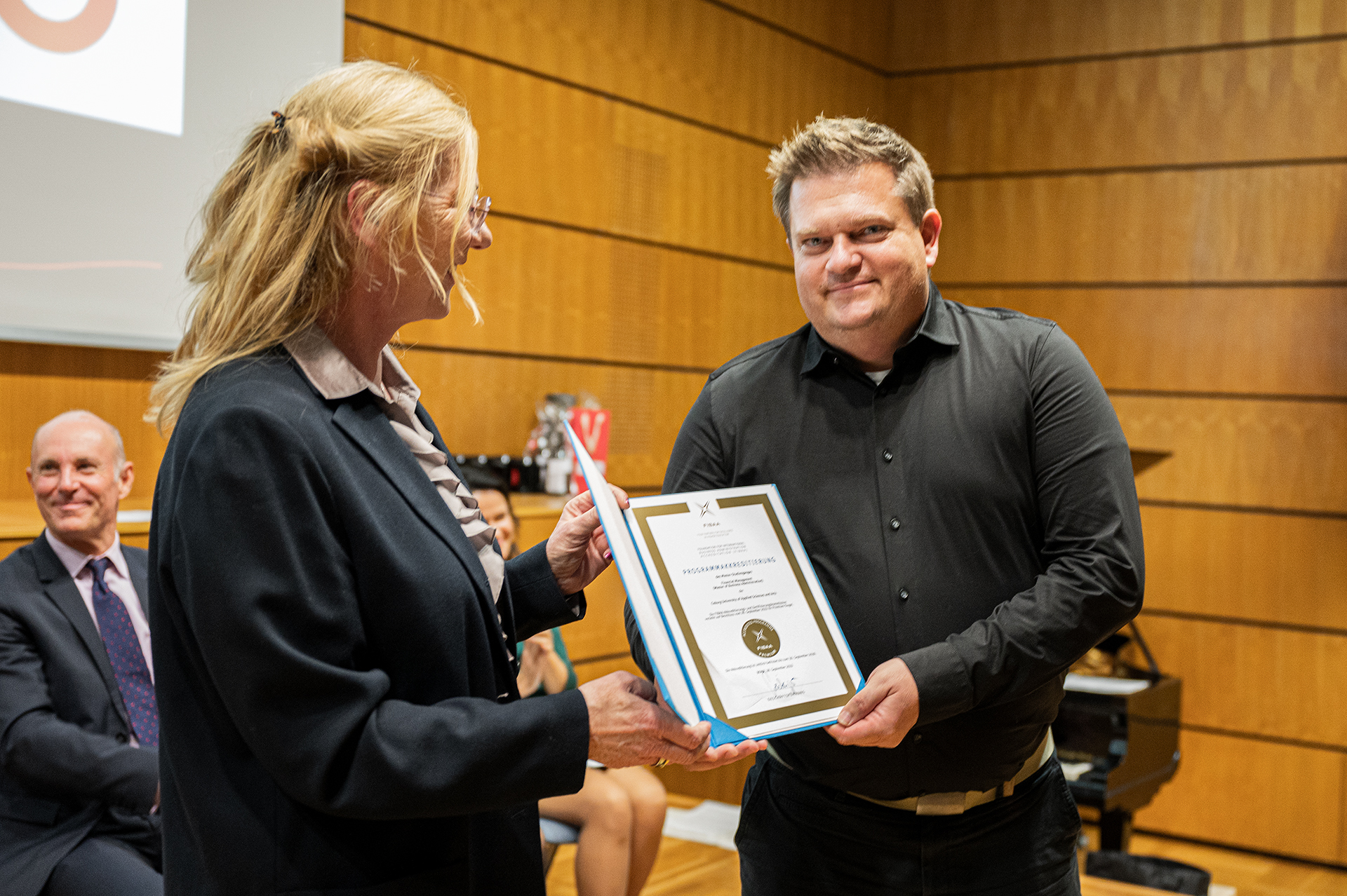 A man in a black shirt receives a framed certificate from a woman with long blonde hair in an indoor setting at Hochschule Coburg. In the background, a seated man in a suit observes the exchange. The wooden walls and visible piano add to the atmosphere of academic achievement.