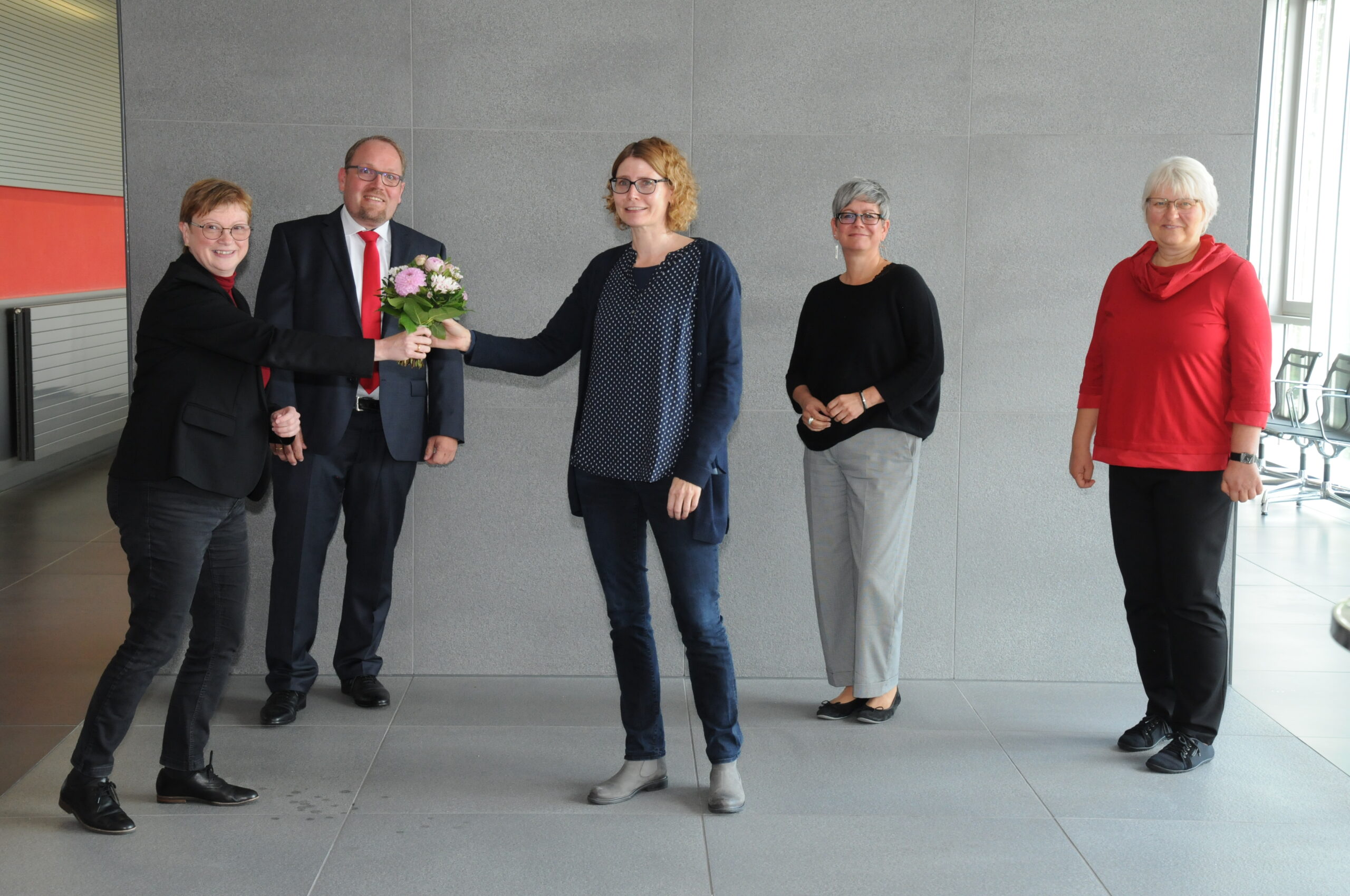 Fünf Menschen stehen gemeinsam vor einer grauen Wand in einem Innenbereich der Hochschule Coburg. Eine Person im schwarzen Outfit überreicht einer anderen Person im blauen Pullover einen Blumenstrauß. Drei weitere, zwei Frauen und ein Mann, lächeln und beobachten die Szene. Im Hintergrund sind Stühle zu sehen.