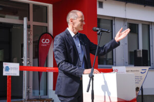 Ein Mann im Anzug steht an einem Rednerpult und spricht in ein Mikrofon. Er gestikuliert mit seiner linken Hand vor dem mit einem Band verzierten Eingang der Hochschule Coburg, eingerahmt von Schildern und Logos, die auf ihre Bedeutung hinweisen.