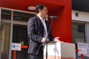 Ein Mann im Anzug spricht in ein Mikrofon an einem Podium vor der roten Wand und den Glastüren der Hochschule Coburg. Schilder und ein rotes Band im Hintergrund deuten auf eine formelle Veranstaltung oder Zeremonie hin, die einen wichtigen Anlass für die Institution markiert.