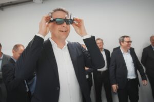 Ein Mann im Anzug, der mit der Hochschule Coburg verbunden ist, lächelt und trägt eine futuristische Virtual-Reality-Brille. Im Hintergrund beobachten mehrere andere Personen in Anzügen aufmerksam und tragen zur professionellen und geschäftlichen Atmosphäre bei.