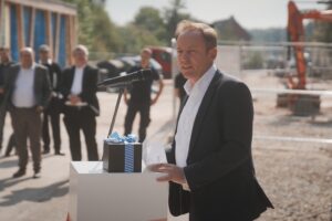 Ein Mann im Anzug spricht an einem sonnigen Tag im Freien an einem Podium mit Mikrofon bei der Veranstaltung der Hochschule Coburg. Auf dem Podium steht eine Geschenkbox mit einer Schleife. Im Hintergrund stehen mehrere Personen neben Baugeräten und Materialien und feiern dieses neue akademische Vorhaben.