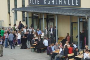 Eine große Gruppe von Menschen hat sich vor einem Gebäude mit dem Schild „Alte Kühlhalle“ in der Nähe der Hochschule Coburg versammelt. Sie mischen sich untereinander, sitzen auf Bänken und stehen um hohe Tische herum. Die Atmosphäre scheint wie eine gesellige Veranstaltung an einem sonnigen Tag.