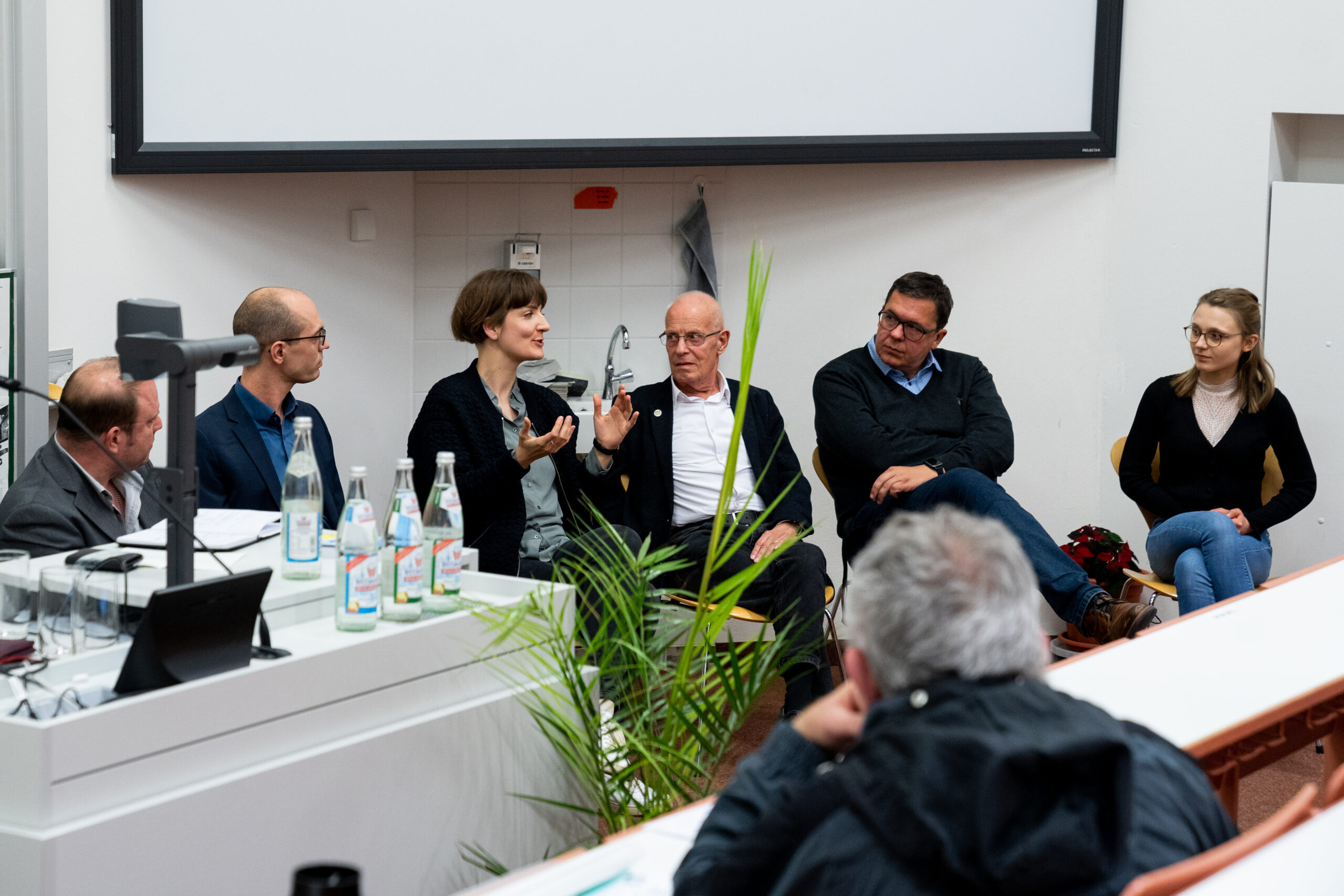 Ein fünfköpfiges Gremium der Hochschule Coburg sitzt vor einem Publikum in einem Hörsaal. Eine Frau gestikuliert beim Sprechen, während die anderen aufmerksam zuhören. Auf einem Tisch in der Nähe stehen Wasserflaschen, daneben ein geöffneter Laptop. Im Vordergrund steht eine Pflanze.