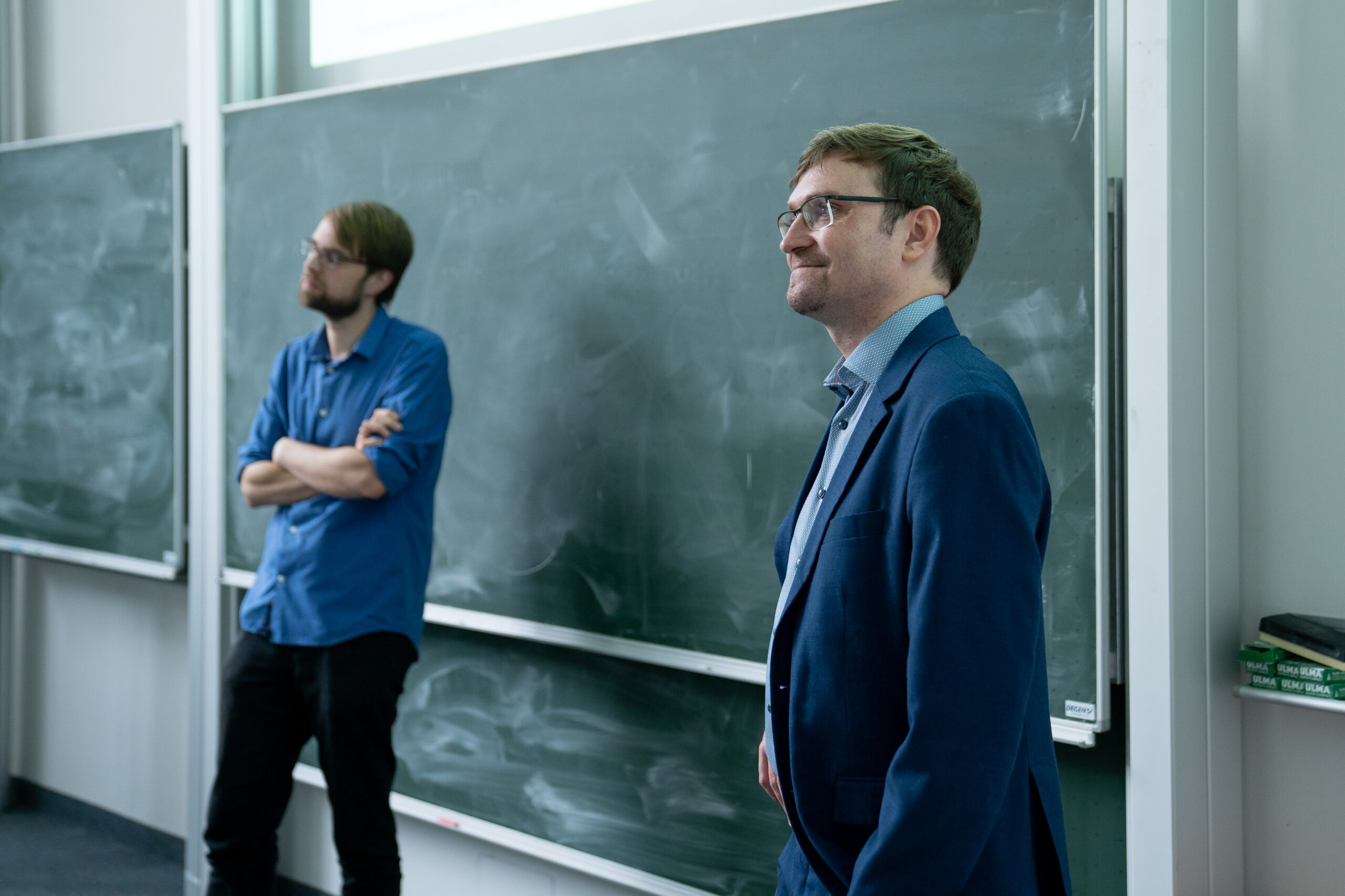 Zwei Männer stehen vor teilweise gelöschten Tafeln, einer in einem blauen Hemd mit verschränkten Armen und der andere in einem blauen Anzug. Die Kulisse, die an ein Klassenzimmer oder einen Hörsaal der Hochschule Coburg erinnert, vermittelt eine akademische Atmosphäre.