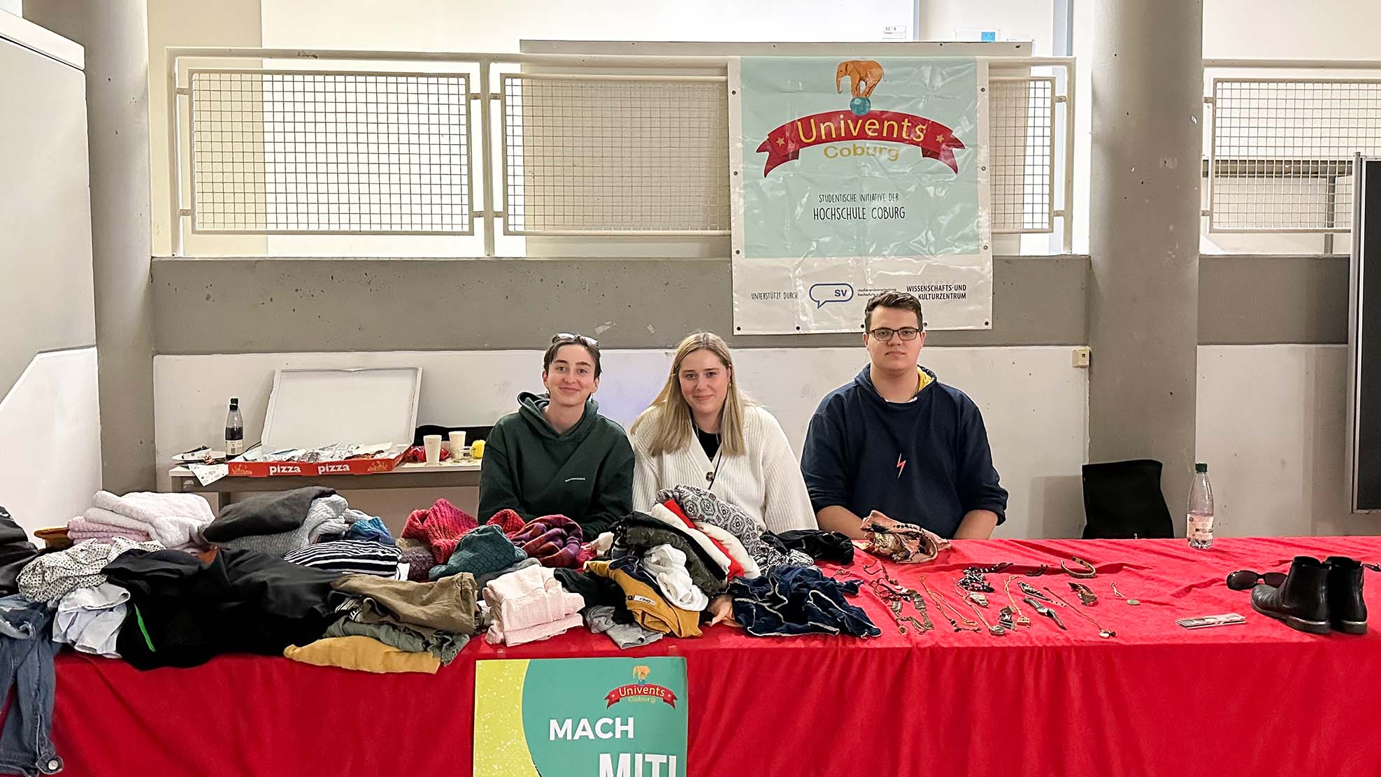 Drei Menschen sitzen hinter einem Tisch auf einem Flohmarkt. Die Tischplatte ist mit verschiedenen Gegenständen bedeckt, darunter Kleidung, Bücher und Schuhe. Über ihnen hängt ein Banner mit der Aufschrift „Univents Coburg“. Davor wirbt ein buntes Plakat mit der Aufschrift „MACH MIT!“ stolz für den Geist der Hochschule Coburg.