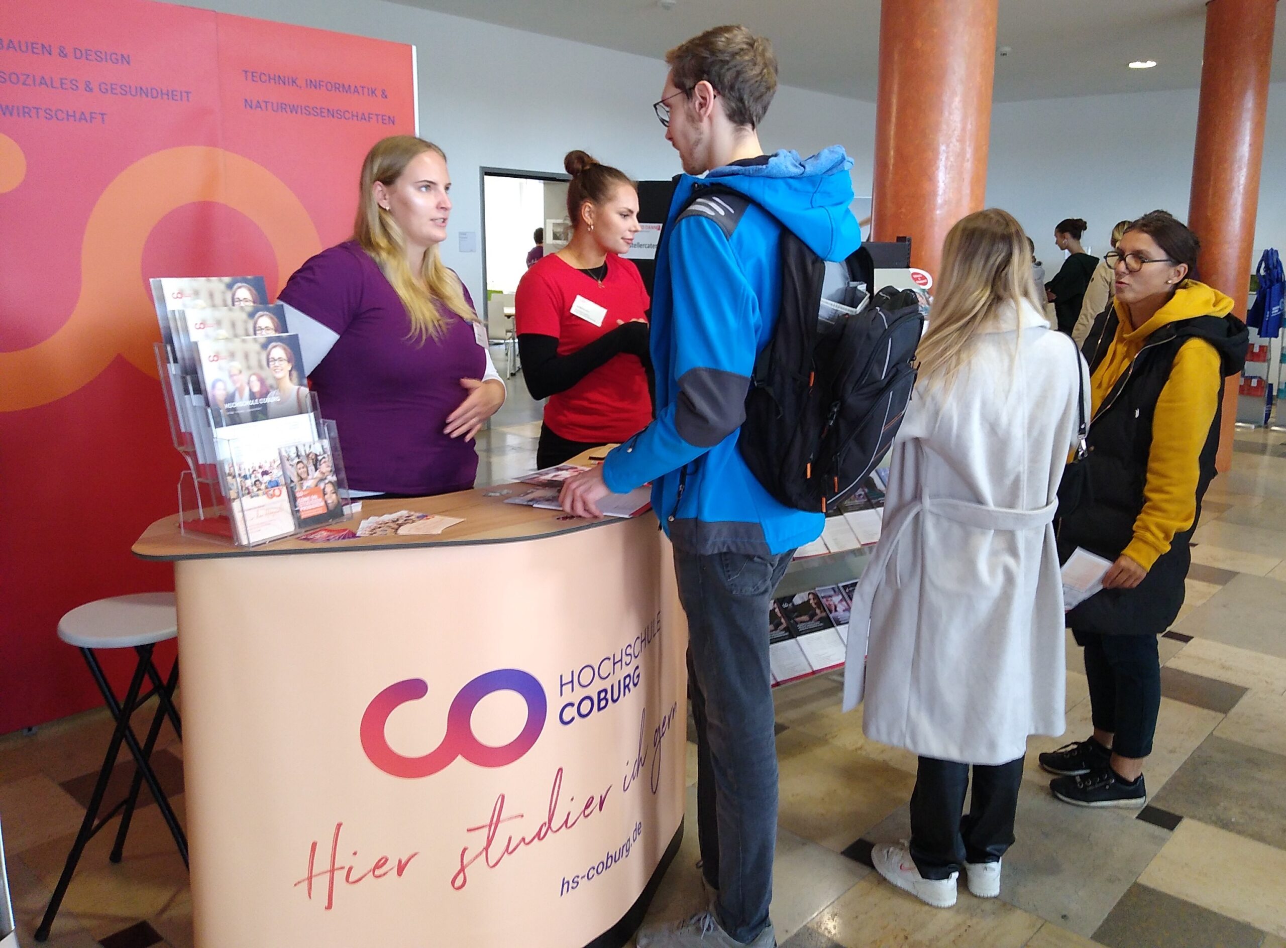 Am Informationsstand der Hochschule Coburg herrscht reges Treiben, während zwei Vertreter mit den Besuchern sprechen. Dahinter informieren Broschüren und eine Schautafel vor einem leuchtend roten und orangefarbenen Hintergrund über die Studienfächer.