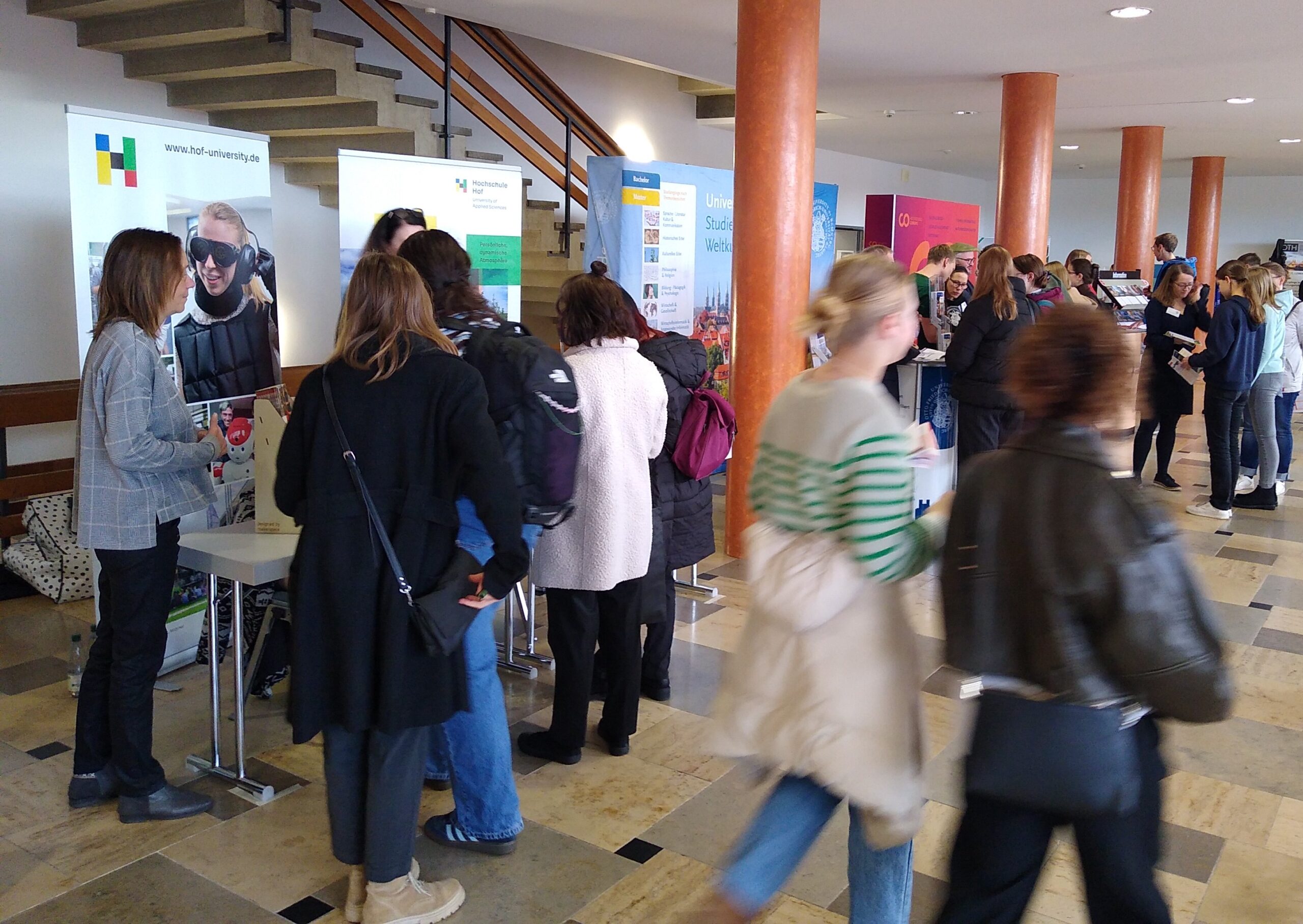 Besucher einer Jobmesse an der Hochschule Coburg sprechen mit Personalvermittlern an verschiedenen Unternehmensständen. Im Hintergrund sind Banner und Werbematerialien zu sehen, während Personen Broschüren in die Hand nehmen und mit Unternehmensvertretern plaudern.