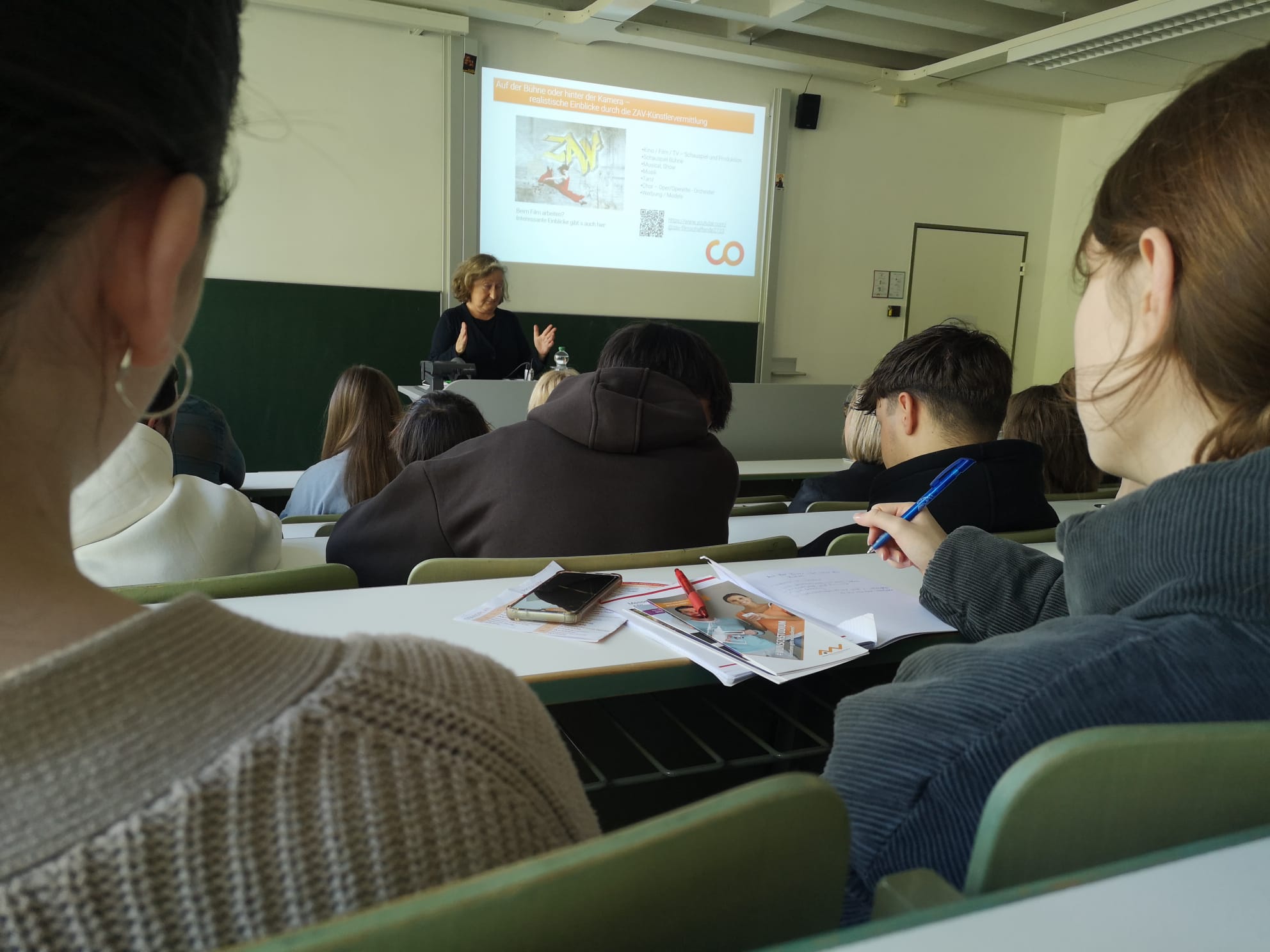 Eine Frau unterrichtet in einem Hörsaal der Hochschule Coburg. Sie steht vor einer Projektionsleinwand, auf der eine Präsentation läuft. Die Studierenden sitzen in Reihen und machen sich fleißig Notizen. Auf einem Tisch im Vordergrund liegen Papiere und ein Telefon, was die akademische Atmosphäre einfängt.