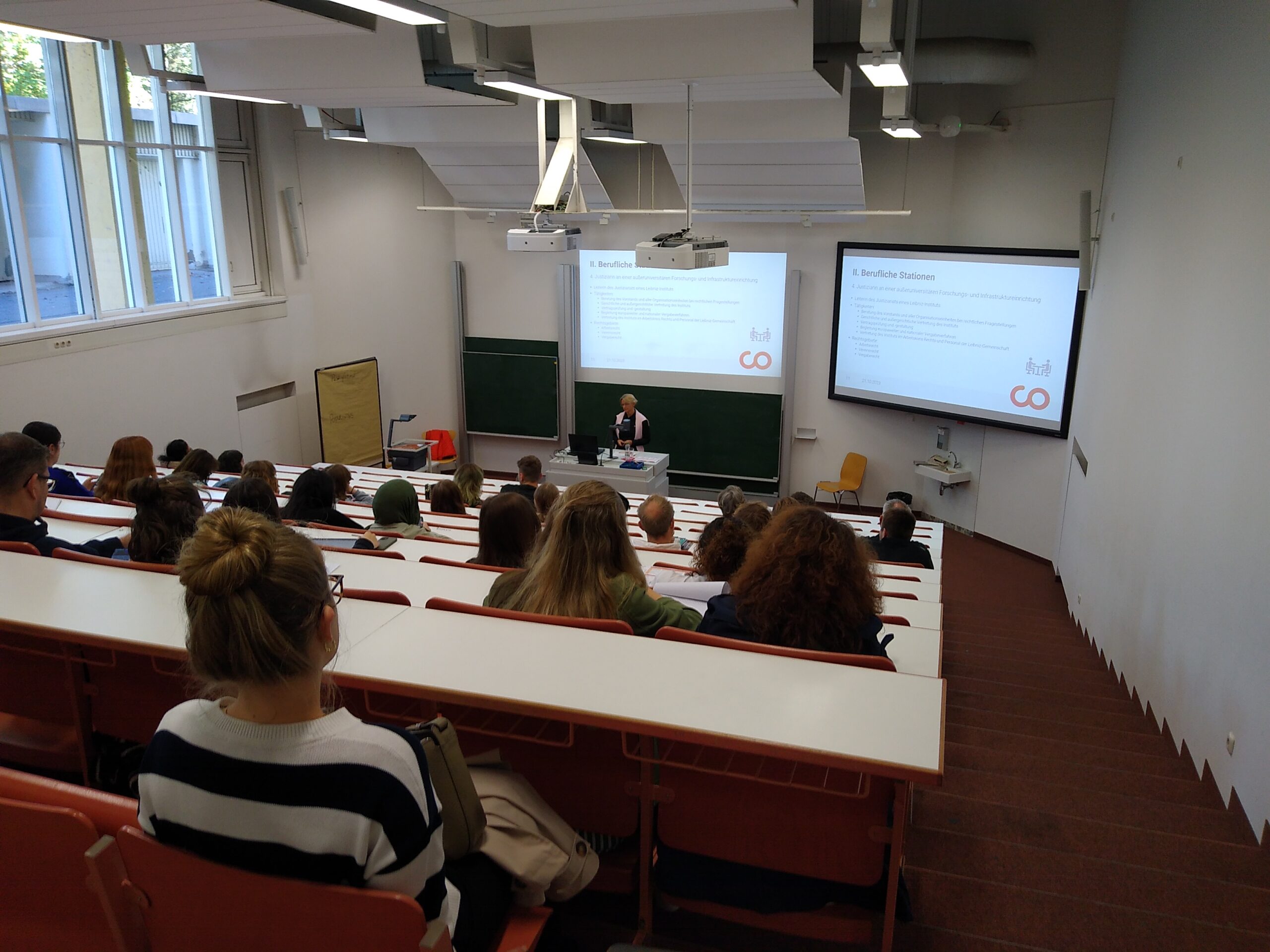 Ein Hörsaal der Hochschule Coburg ist voller Studenten, die auf roten Stühlen sitzen und einem Vortragenden in der ersten Reihe aufmerksam zuhören. Auf zwei großen Bildschirmen wird eine Präsentation gezeigt, während durch die Fenster auf der linken Seite des Raums Tageslicht hereinfällt.