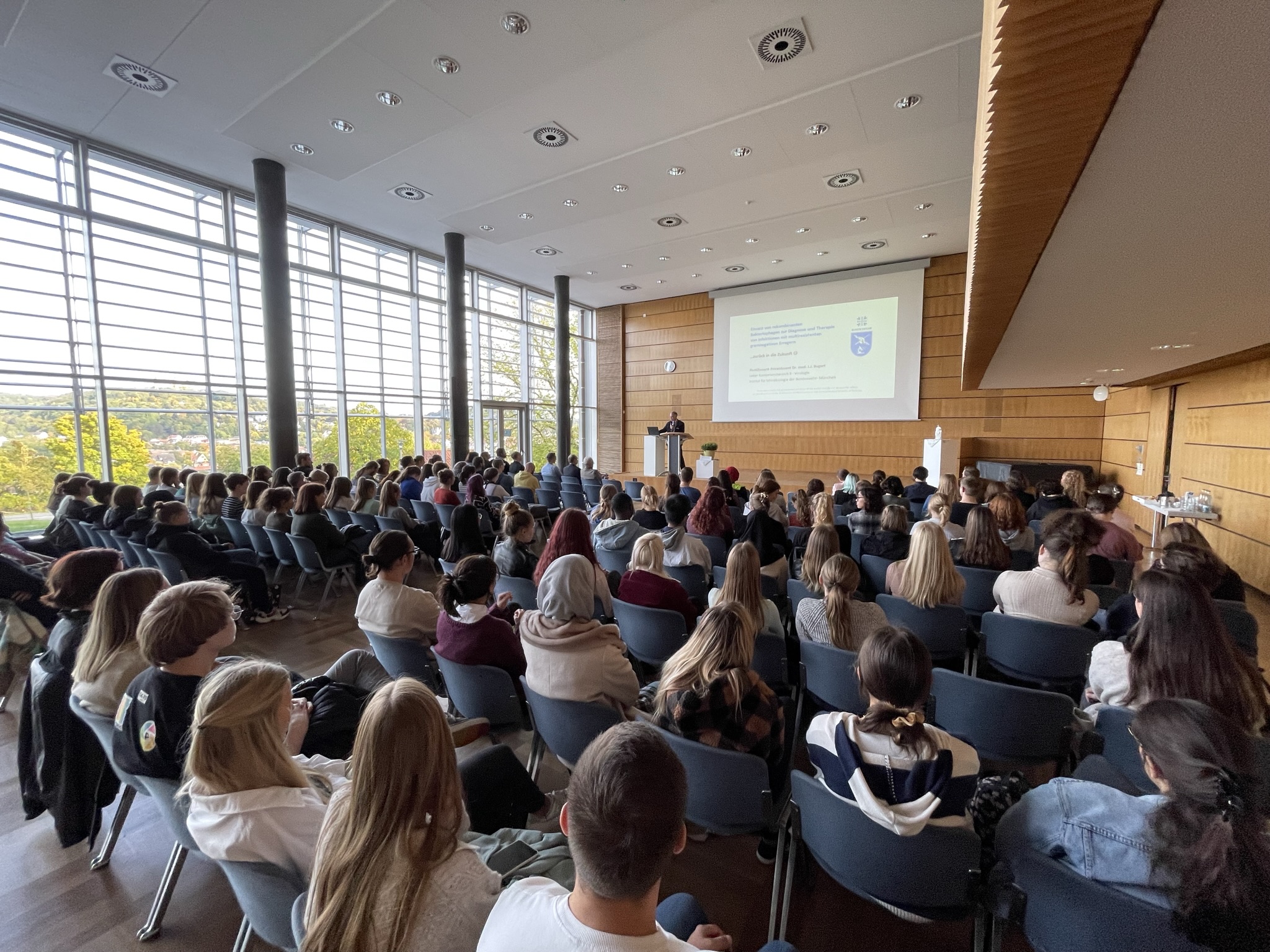 Eine große Gruppe von Menschen sitzt in einem modernen Konferenzraum mit Holzvertäfelung und großen Fenstern der Hochschule Coburg und hört aufmerksam einer Präsentation zu, die auf eine Leinwand an der Vorderseite des Raumes projiziert wird.