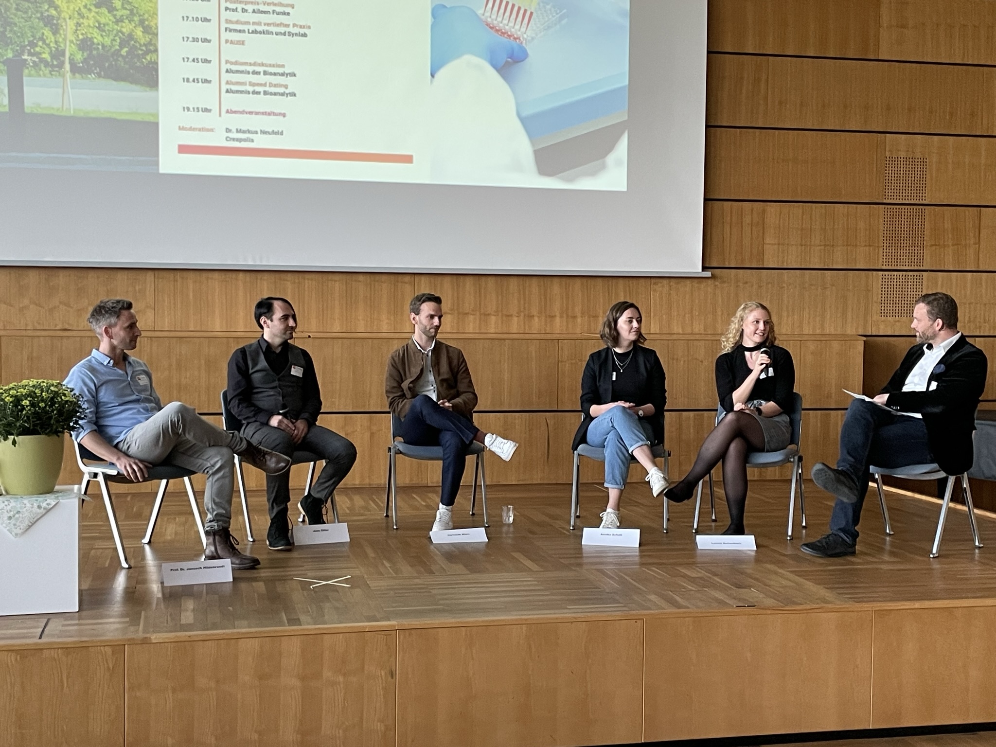 Sechs Personen sitzen bei einer Podiumsdiskussion an der Hochschule Coburg auf einer Bühne. Fünf sitzen auf Stühlen mit Blick zum Publikum, einer spricht in ein Mikrofon. Hinter ihnen zeigt ein Bildschirm den Zeitplan, seitlich der Bühne steht eine Topfpflanze.