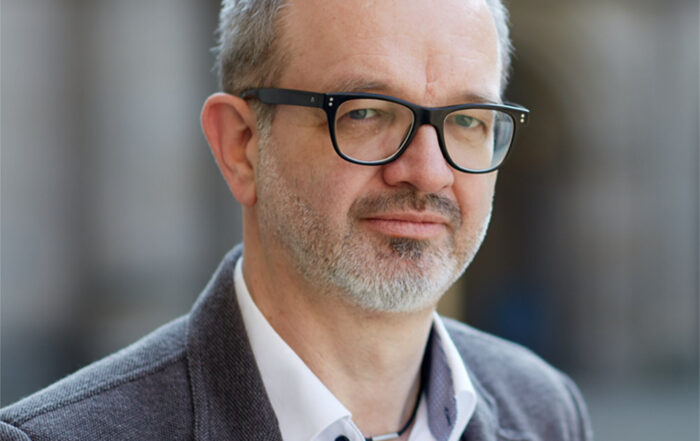 Ein Mann mit kurzen grauen Haaren und Brille, der eine graue Jacke, ein weißes Hemd und eine Weste trägt, steht im Freien vor einem unscharfen Hintergrund. Sein gelassenes Auftreten spiegelt seine Verbindung zur Hochschule Coburg wider.