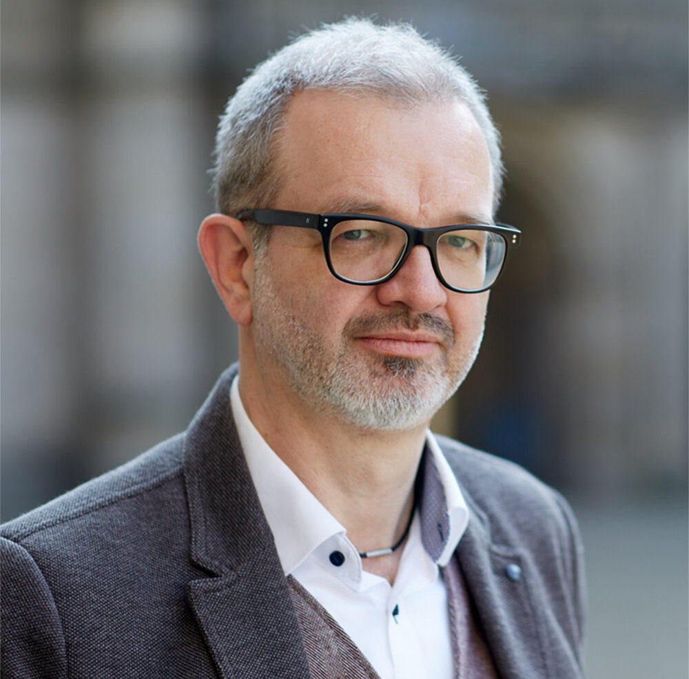 Ein Mann mit kurzen grauen Haaren und Brille, der eine graue Jacke, ein weißes Hemd und eine Weste trägt, steht im Freien vor einem unscharfen Hintergrund. Sein gelassenes Auftreten spiegelt seine Verbindung zur Hochschule Coburg wider.