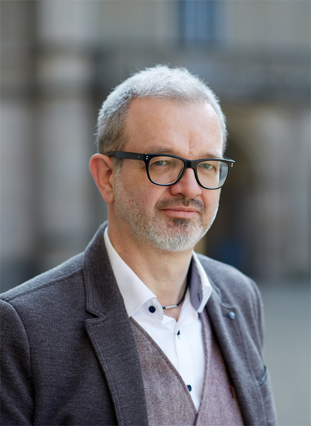 Ein Mann mit kurzen grauen Haaren und Brille, der eine graue Jacke, ein weißes Hemd und eine Weste trägt, steht im Freien vor einem unscharfen Hintergrund. Sein gelassenes Auftreten spiegelt seine Verbindung zur Hochschule Coburg wider.