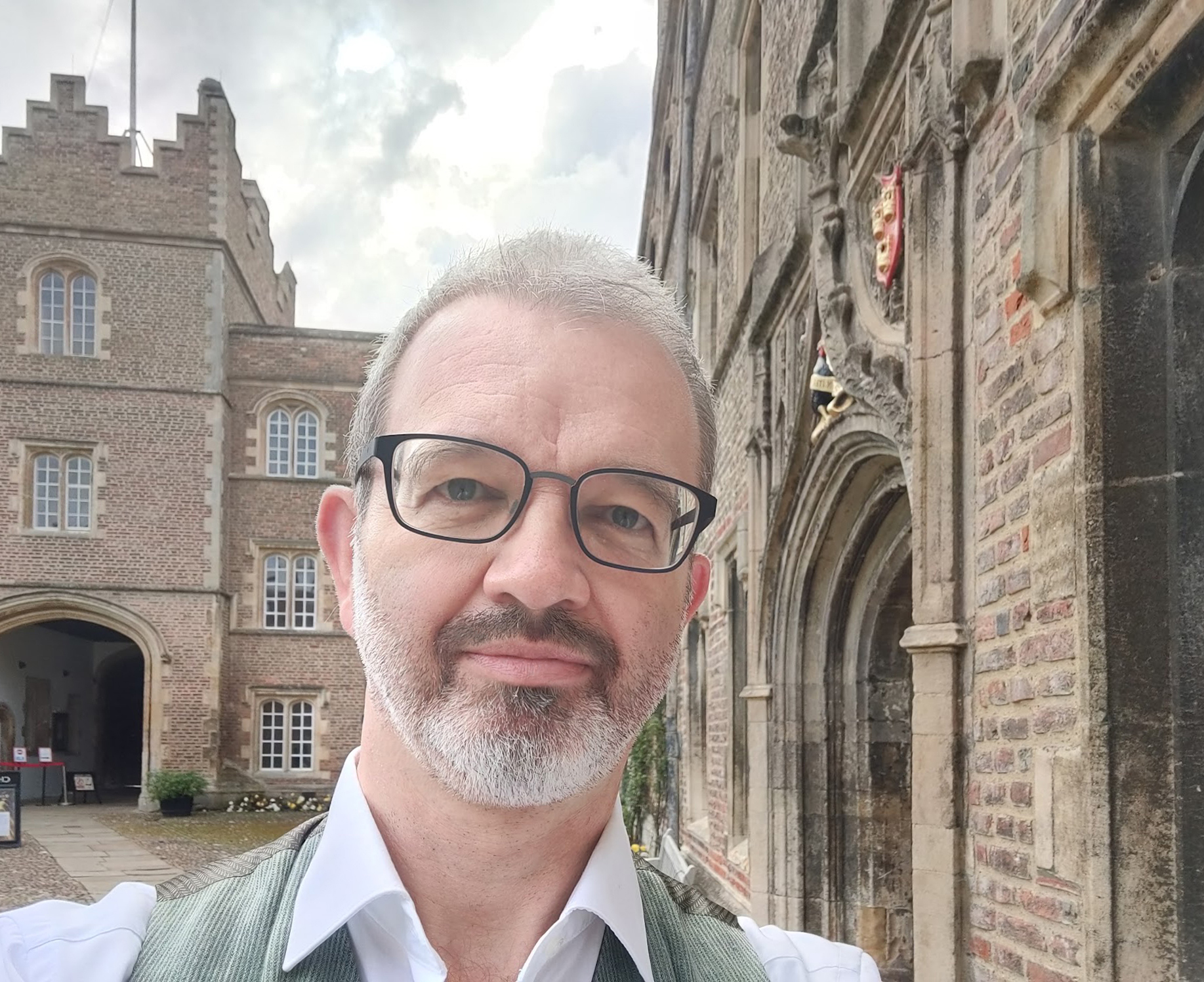 Ein Mann mit Brille und Bart macht ein Selfie vor der Hochschule Coburg. Das Foto zeigt das historische Backsteingebäude mit den gewölbten Türen und dem Wappen. Der Himmel ist leicht bewölkt.