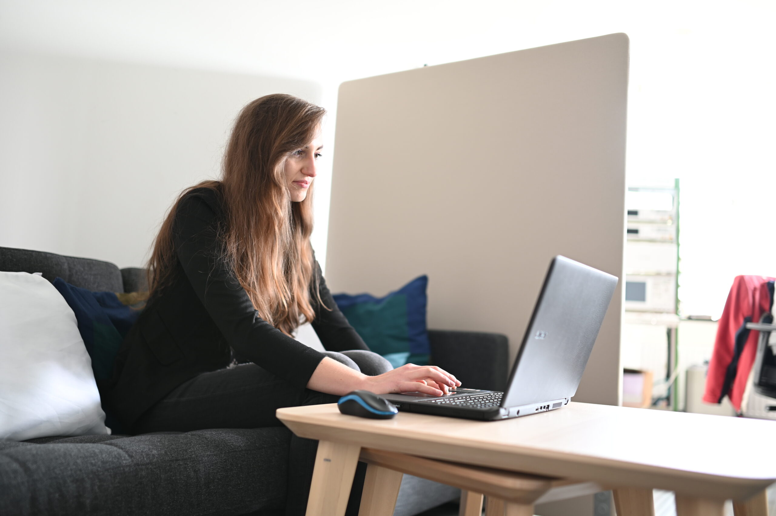 Eine Frau mit langen Haaren sitzt auf einem Sofa und tippt auf einem Laptop, der auf einem Holztisch steht. Sie arbeitet wahrscheinlich an Aufgaben für die Hochschule Coburg. Eine Computermaus liegt daneben. Der Raum ist sanft beleuchtet, im Hintergrund ist ein Bildschirm als Raumteiler zu sehen, auf dem Sofa liegen Kissen.