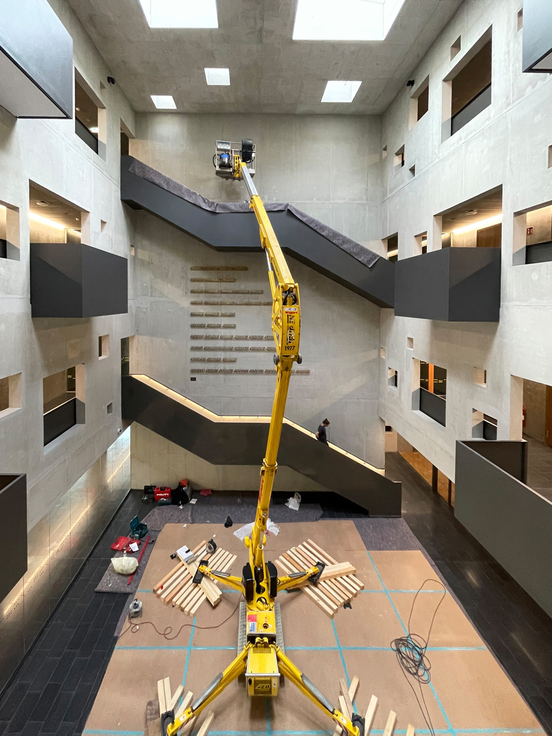 Im modernen Innenraum des Hochhauses der Hochschule Coburg steht ein Bauaufzug mit gelbem Arm, umgeben von Betonwänden und zahlreichen Fensteröffnungen. Arbeiter sind damit beschäftigt, den Aufzug einzustellen, während auf dem Boden Baumaterialien verstreut liegen.