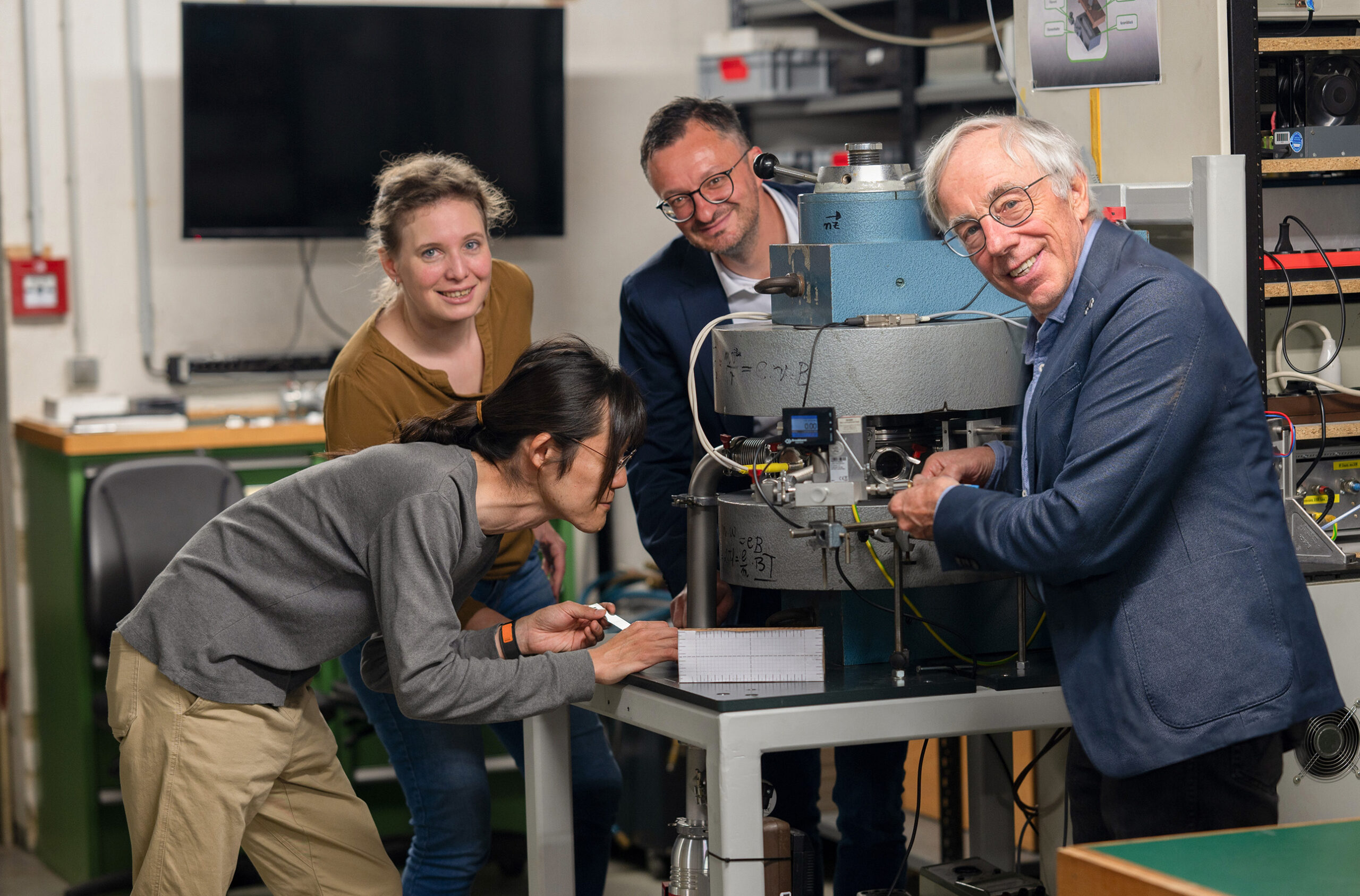 In einem Labor der Hochschule Coburg arbeiten vier Personen gemeinsam an einem wissenschaftlichen Instrument. Einer schaut sich das Gerät genau an, während andere zuschauen und assistieren. Der Raum ist voll mit technischen Geräten und einem Computermonitor und spiegelt den Innovationsgeist dieser renommierten Institution wider.