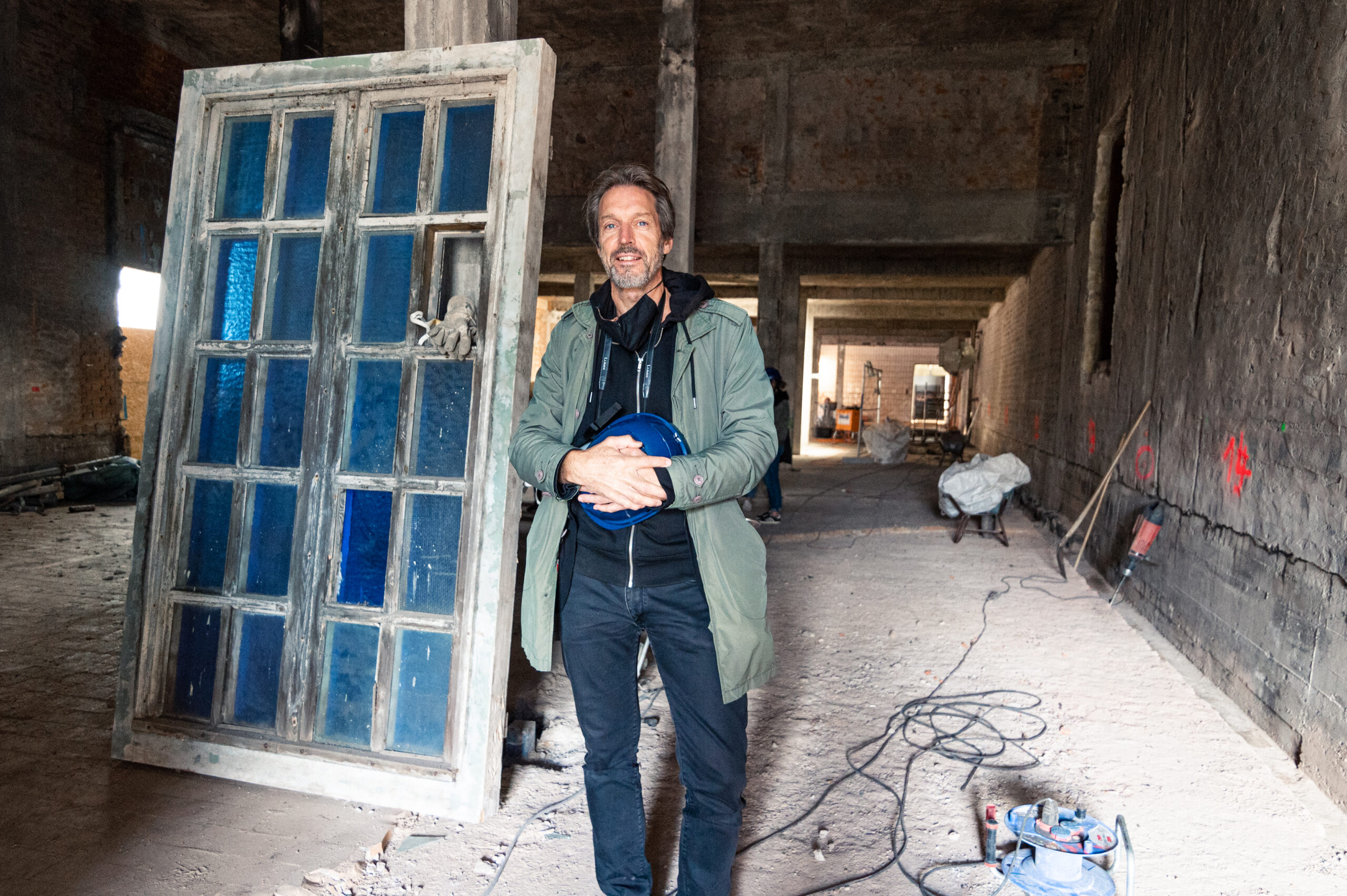 Ein Mann in einer grünen Jacke steht in einem Rohbau der Hochschule Coburg und hält eine blaue Kabeltrommel in der Hand. Hinter ihm befindet sich eine große, verwitterte Tür mit blauen Glasscheiben. Überall im Raum liegen Baumaterialien und Werkzeuge verstreut.