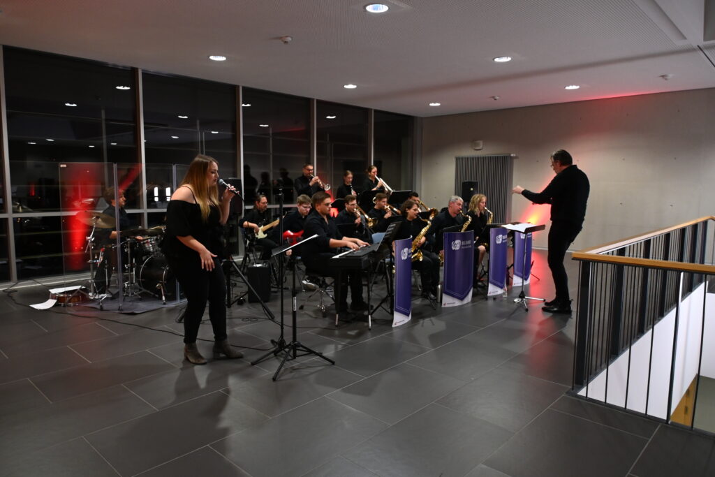 A jazz band from Hochschule Coburg performs indoors with a female vocalist. The conductor leads the ensemble, which includes a pianist, brass players, and a drummer. The modern setting features large windows and dim lighting, creating an intimate ambiance for the unseen audience.