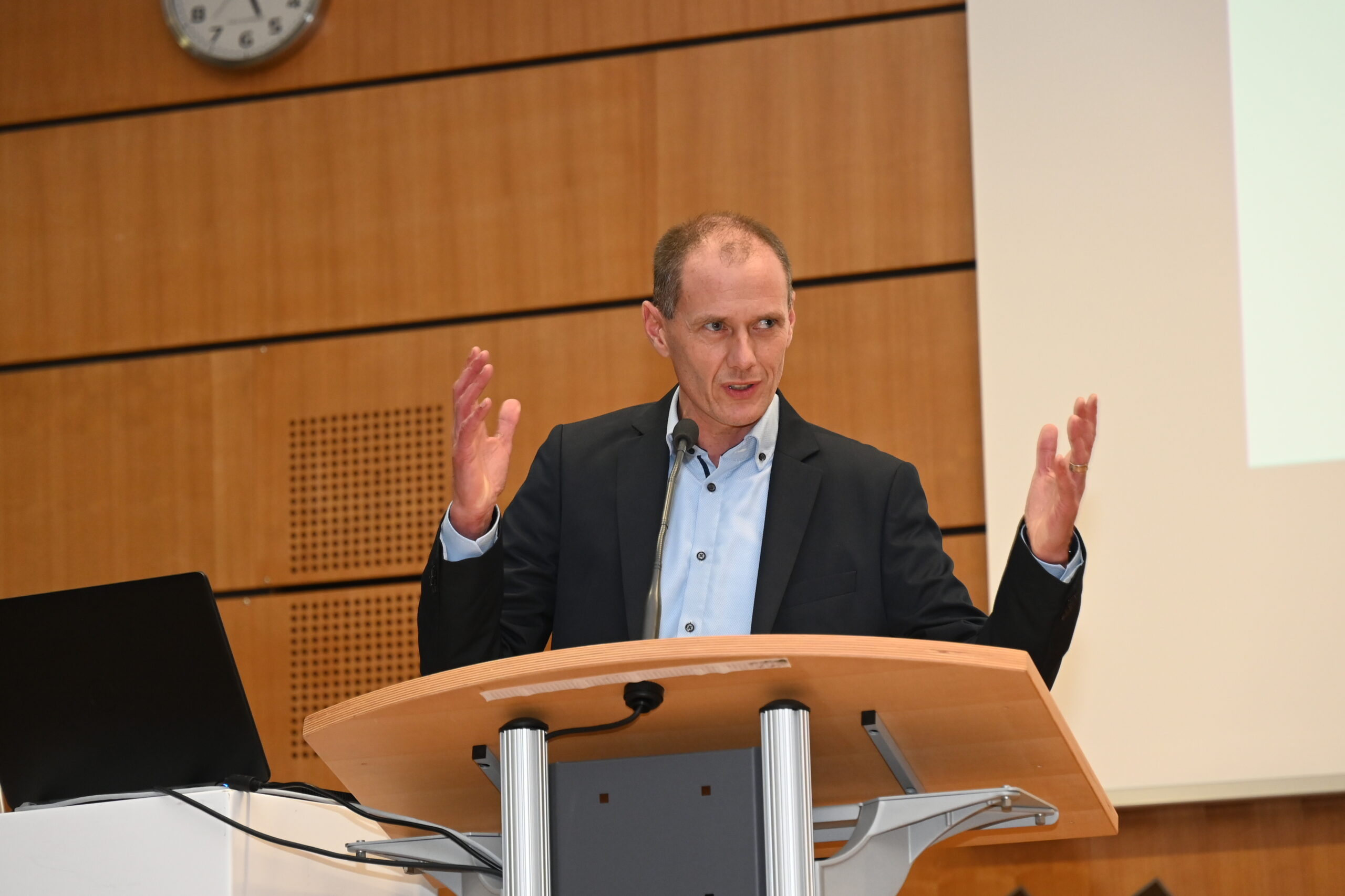 Eine Person steht an einem Holzpodest mit dem Emblem der Hochschule Coburg und gestikuliert mit beiden Händen. Sie trägt einen dunklen Blazer und ein hellblaues Hemd. Auf dem Podest steht ein Laptop, im Hintergrund ist eine Wanduhr zu sehen.