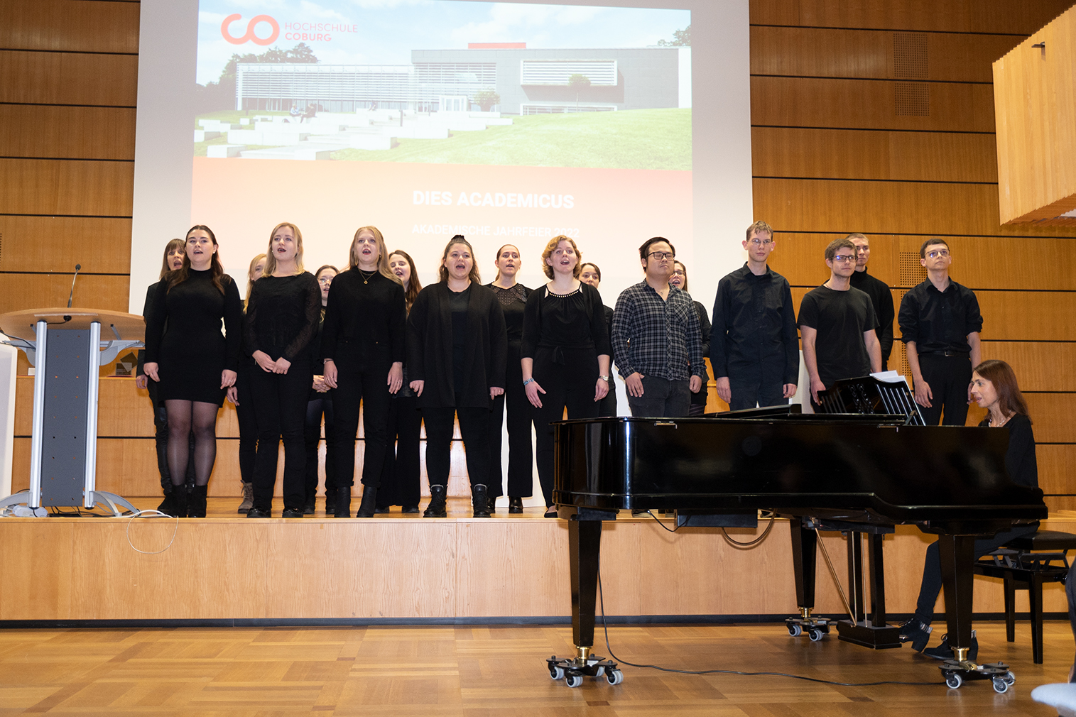 Ein Chor aus zwölf schwarz gekleideten Personen tritt auf der Bühne neben einem Pianisten an einem Flügel auf. Im Hintergrund läuft auf einer Präsentationsleinwand „Dies Academicus“, während sich die Hochschule Coburg in einem holzgetäfelten Hörsaal versammelt.