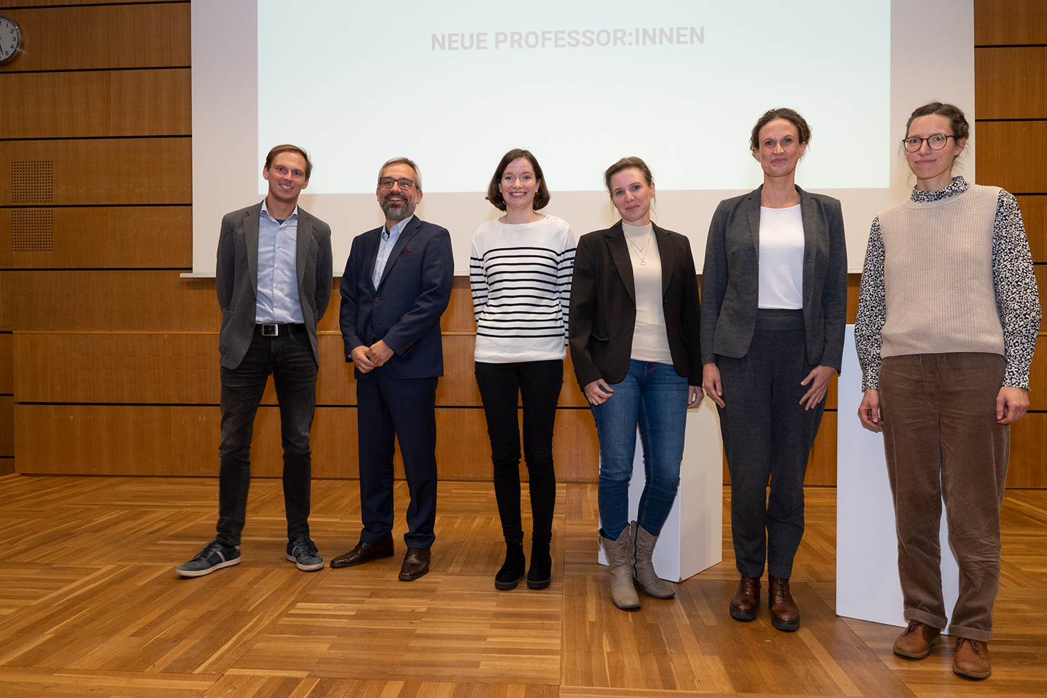 Eine Gruppe von sechs Personen steht in einer Reihe auf einem Holzboden und lächelt in die Kamera. Sie befinden sich in einem Raum mit holzgetäfelten Wänden und einer Projektionsleinwand, auf der im Hintergrund die Worte „Neue Professor:innen“ zu sehen sind. Ein inspirierender Moment an der Hochschule Coburg.