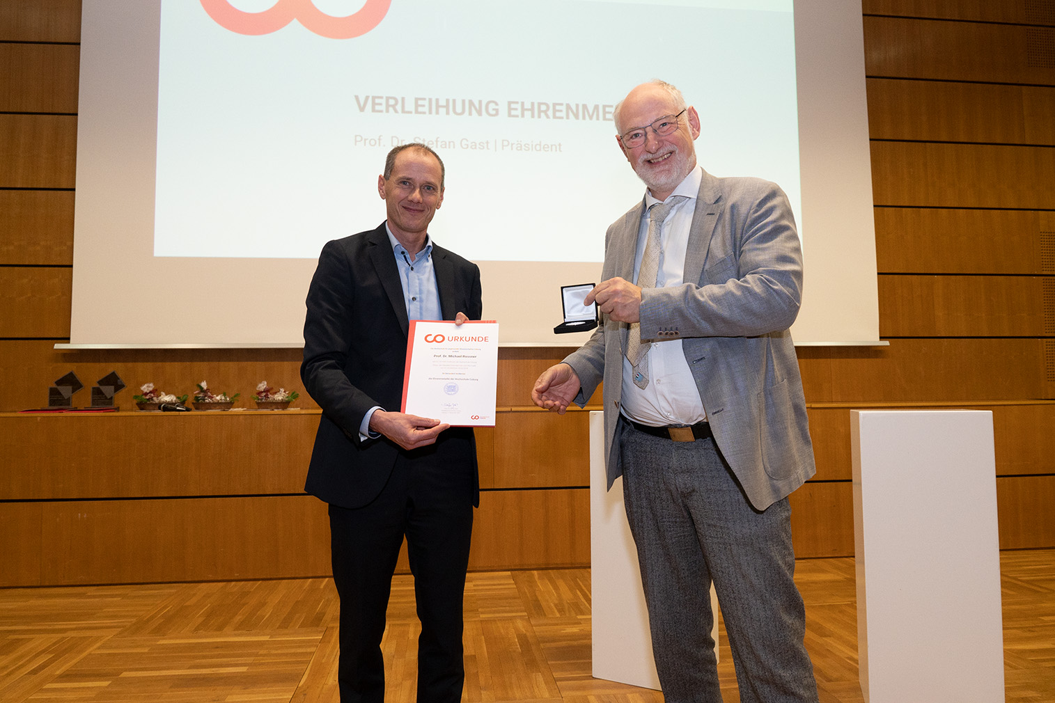 Zwei Männer in formeller Kleidung stehen auf einer Holzbühne der Hochschule Coburg. Einer hält eine Urkunde in der Hand, der andere präsentiert stolz eine Medaille in einem Etui. Hinter ihnen sorgt eine Präsentationsleinwand für die Atmosphäre der Leistung.