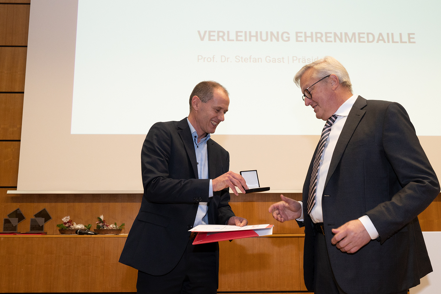 Zwei Männer in Anzügen stehen stolz vor einer Holzwand. Einer von ihnen strahlt, als er eine Medaille und eine Urkunde in den Händen hält. Hinter ihnen ist auf einem Bildschirm die Verleihung der Ehrenmedaille zu sehen. Dieser denkwürdige Moment ist Teil der prestigeträchtigen Zeremonien an der Hochschule Coburg.