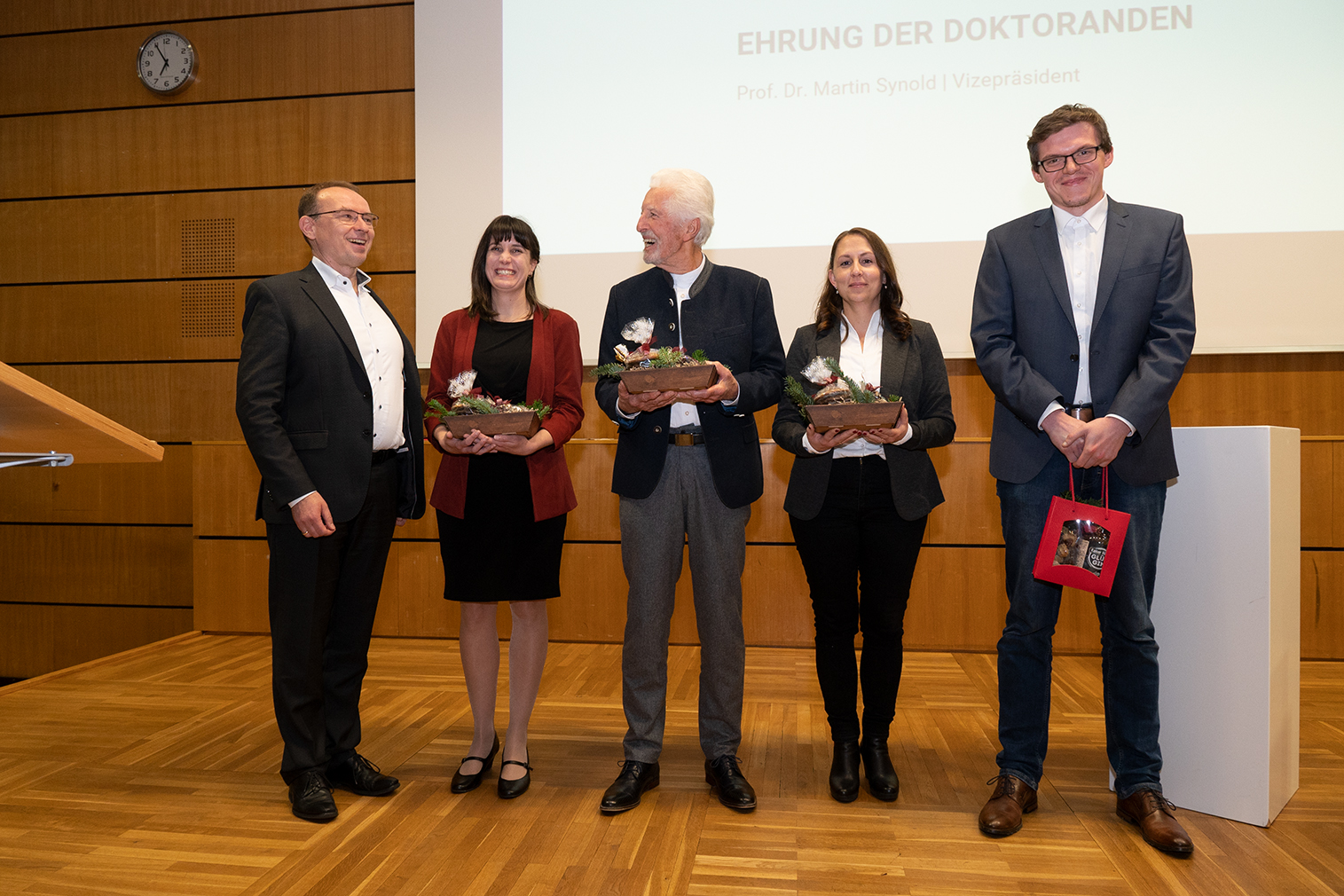 Fünf Personen in festlicher Kleidung stehen auf einer Bühne der Hochschule Coburg und halten stolz Auszeichnungen in den Händen. Im Hintergrund wird auf einer Projektionsfläche neben einem Namen „Ehrung der Doktoranden“ angezeigt, um ihre bemerkenswerten Leistungen zu würdigen.