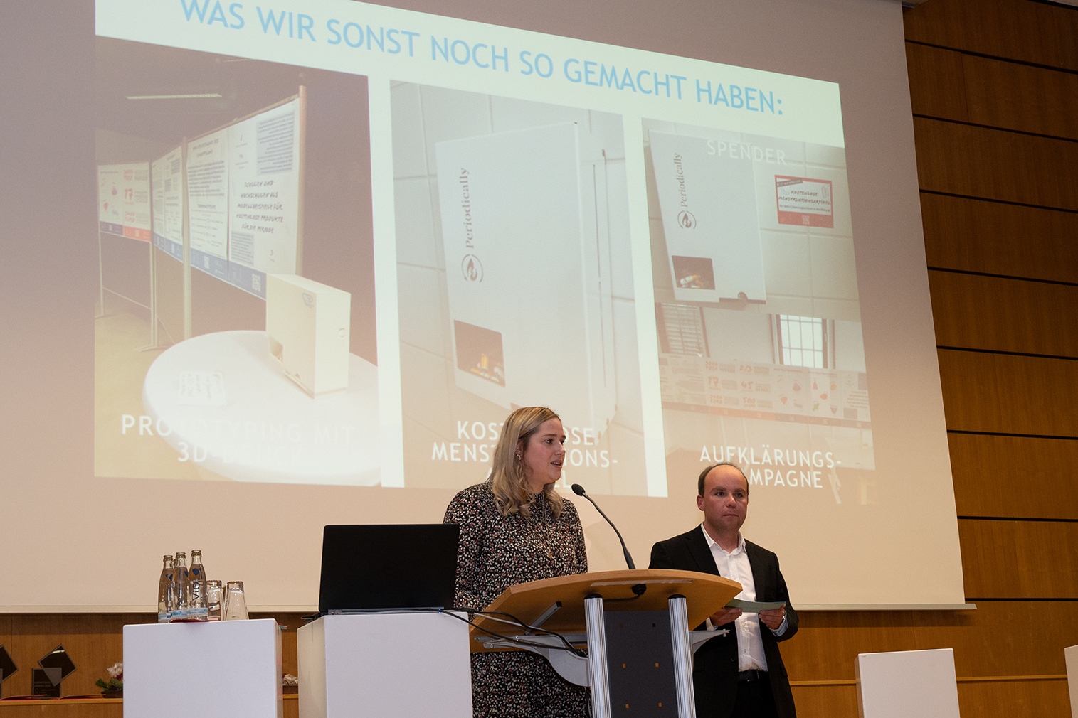 Zwei Menschen stehen an einem Podium und halten einen Vortrag an der Hochschule Coburg. Dahinter hängt eine große Folie mit dem Titel „WAS WIR SONST NOCH SO GEMACHT HABEN“, auf der drei Projekte vorgestellt werden. Der Raum mit seiner Holzvertäfelung strahlt eine moderne, professionelle Atmosphäre aus.