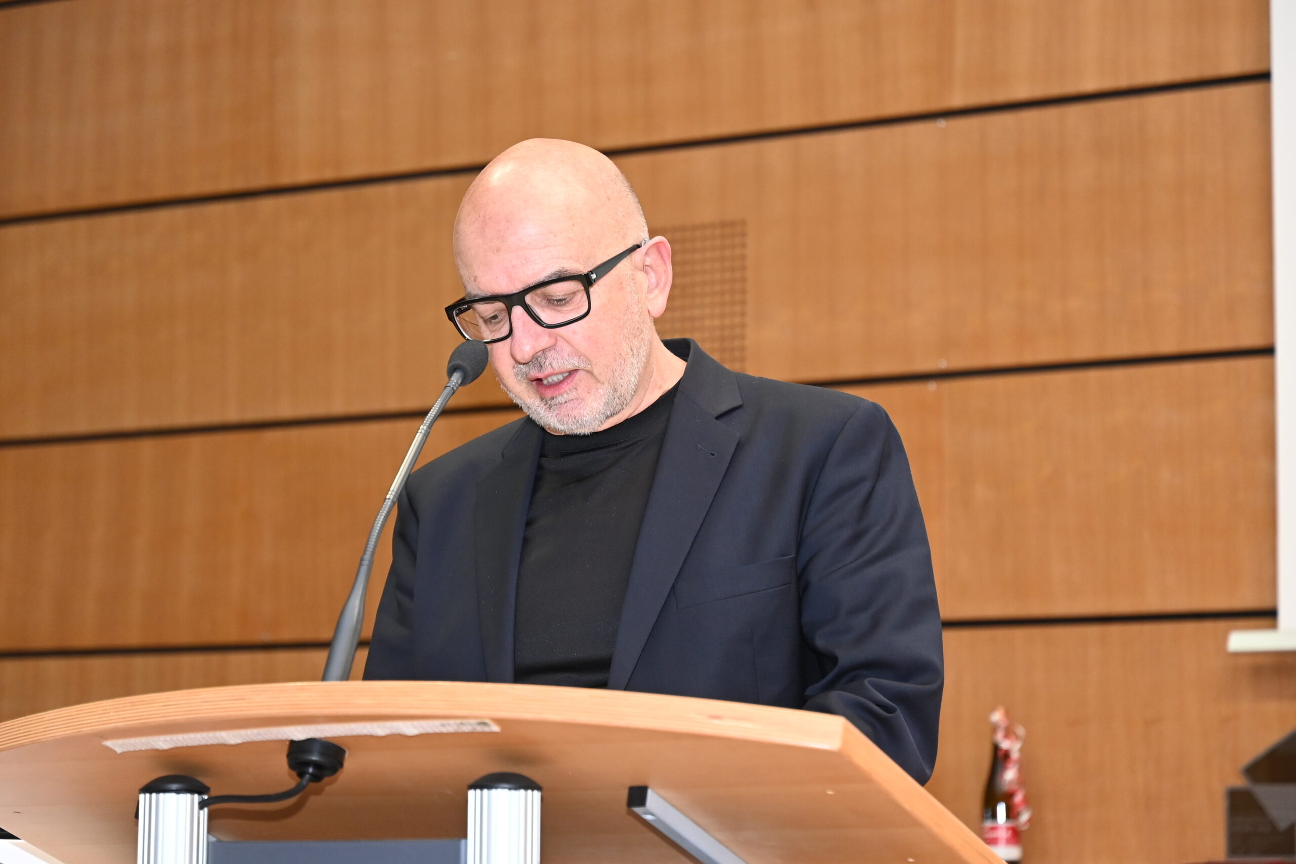 Eine Person mit Brille, rasiertem Kopf und schwarzem Anzug spricht an einem hölzernen Rednerpult vor der holzgetäfelten Wand der Hochschule Coburg in ein Mikrofon.