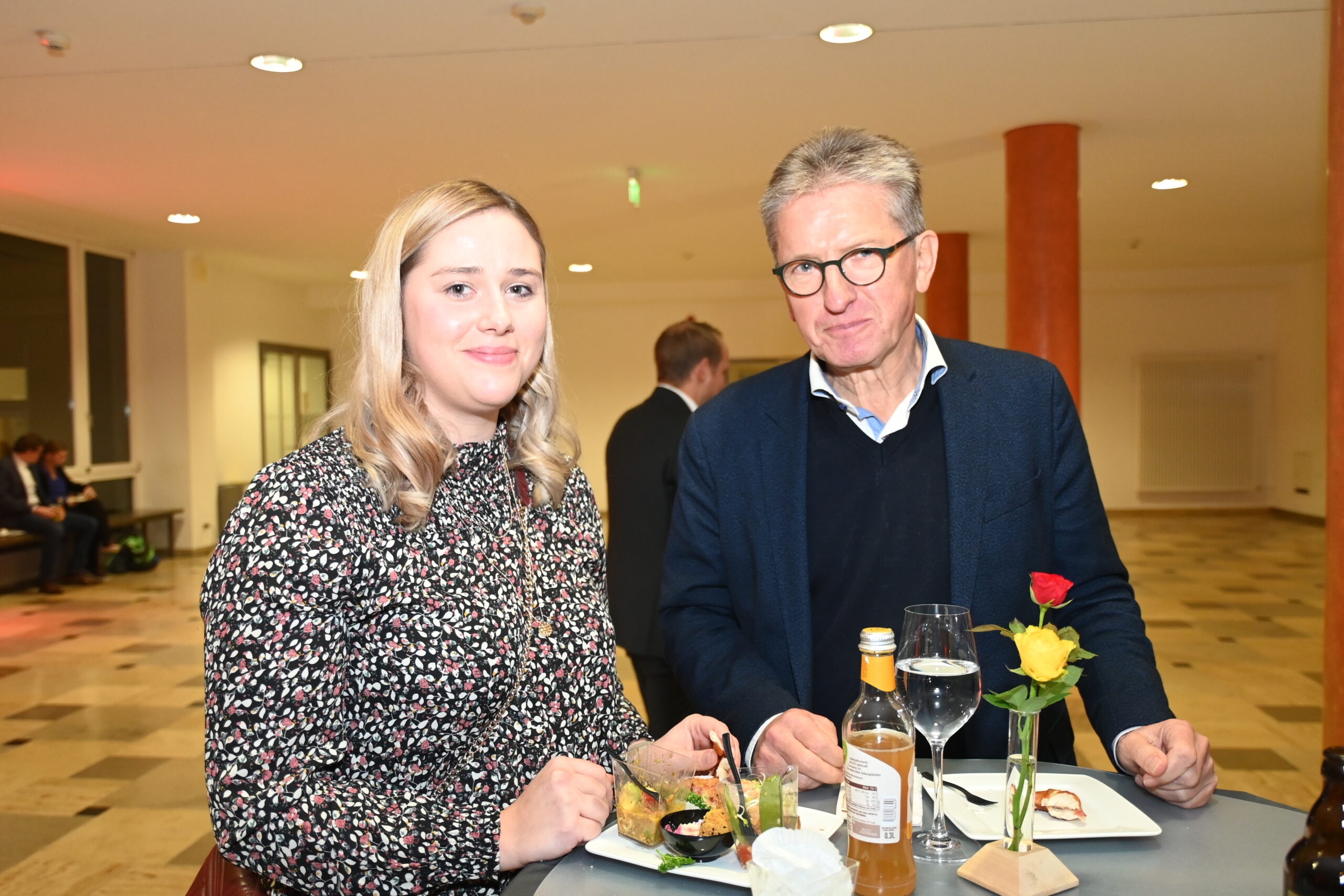 Eine Frau und ein Mann stehen an einem Tisch mit Essen und Getränken in einem hell erleuchteten Raum der Hochschule Coburg. Die Frau trägt eine gemusterte Bluse, während der Mann eine Brille zu dunklem Pullover und Jacke trägt. Eine Rose in einer Vase ergänzt das Ambiente neben edlem Glasgeschirr.