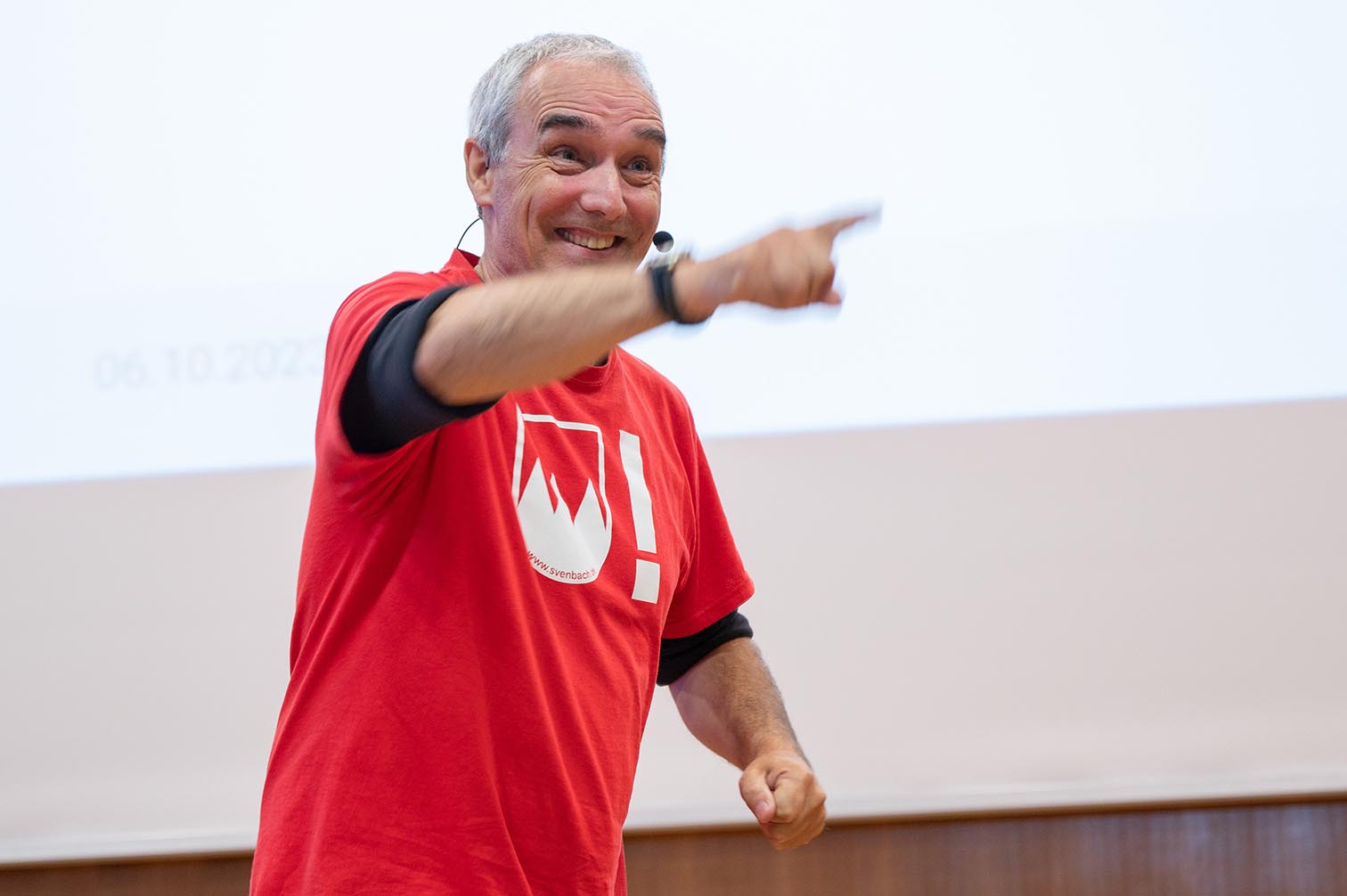 Ein Mann mit kurzen grauen Haaren und einem roten T-Shirt mit weißem Logo spricht in ein Headset-Mikrofon. Er strahlt, zeigt nach vorne und steht selbstbewusst vor einer weißen Leinwand in der Hochschule Coburg.