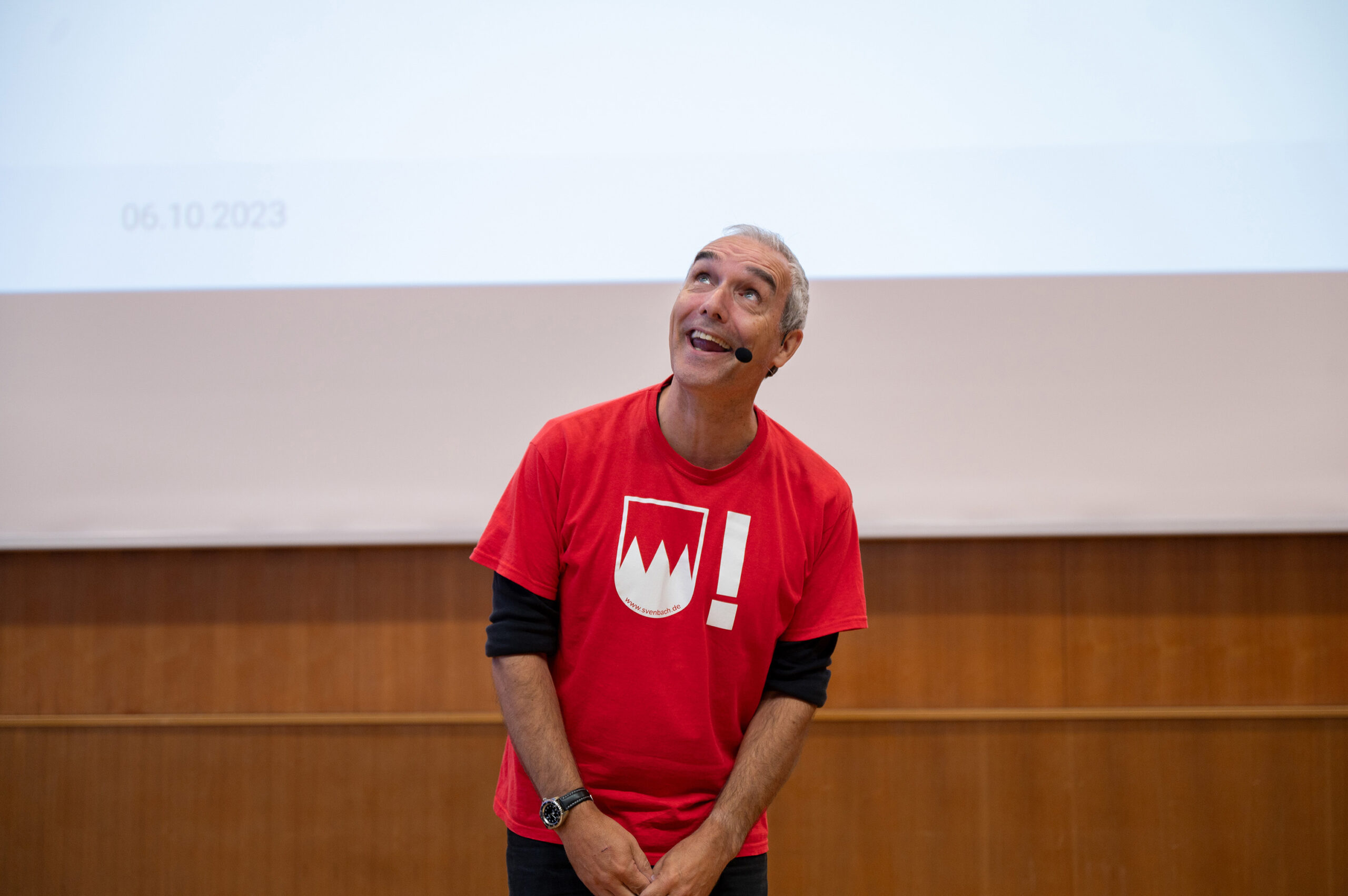 Eine Person in einem roten T-Shirt mit der Aufschrift „Hochschule Coburg“ trägt ein Mikrofon-Headset, blickt nach oben und lächelt. Sie steht vor einer Holztafel und einem weißen Bildschirm, auf dem ein Datum angezeigt wird.