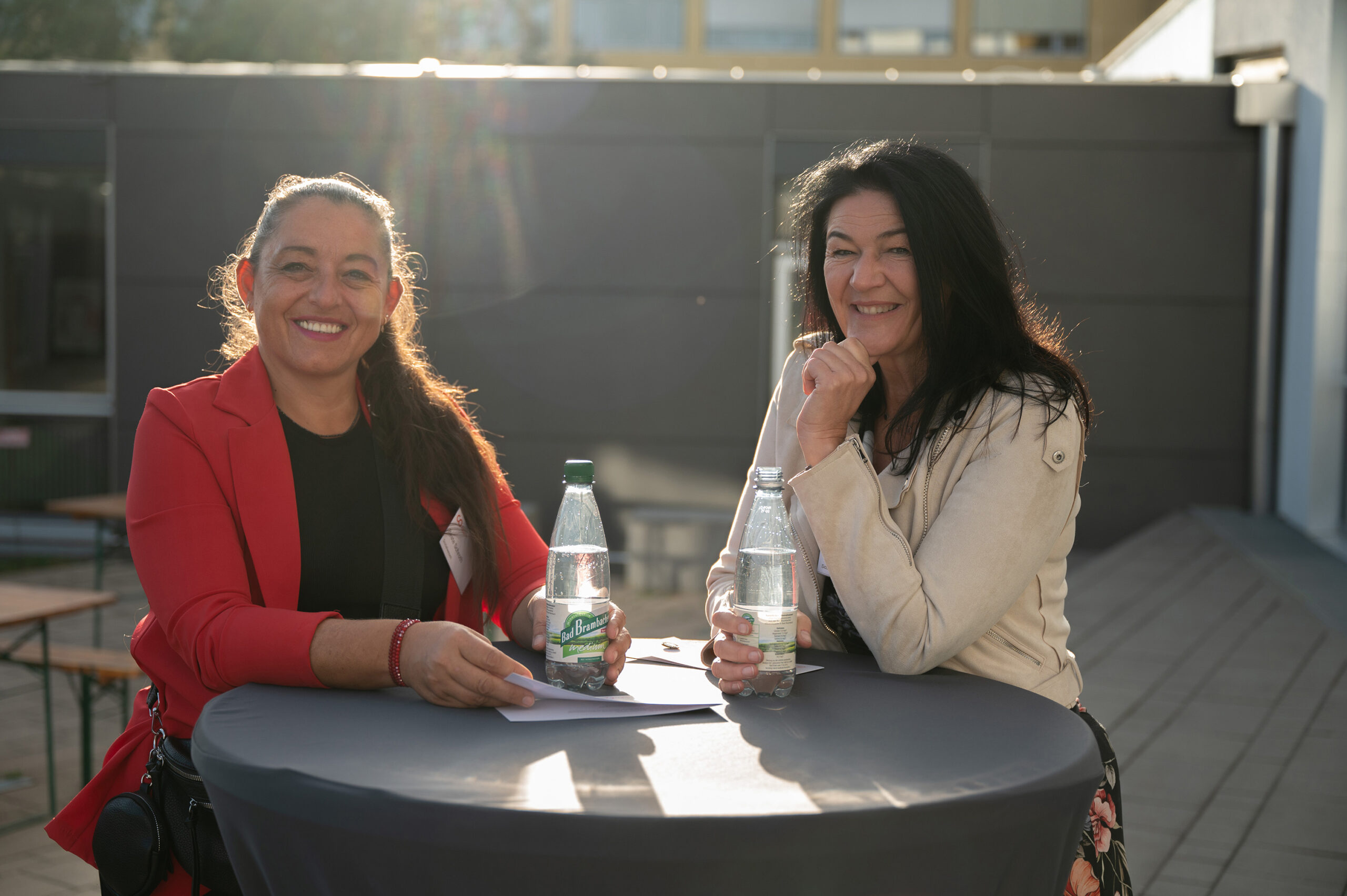 Zwei Frauen sitzen an einem runden Tisch im Freien der Hochschule Coburg und lächeln in die Kamera. Jede hält eine Flasche Mineralwasser in der Hand. Die Sonne scheint hell und sorgt für eine warme und heitere Atmosphäre.