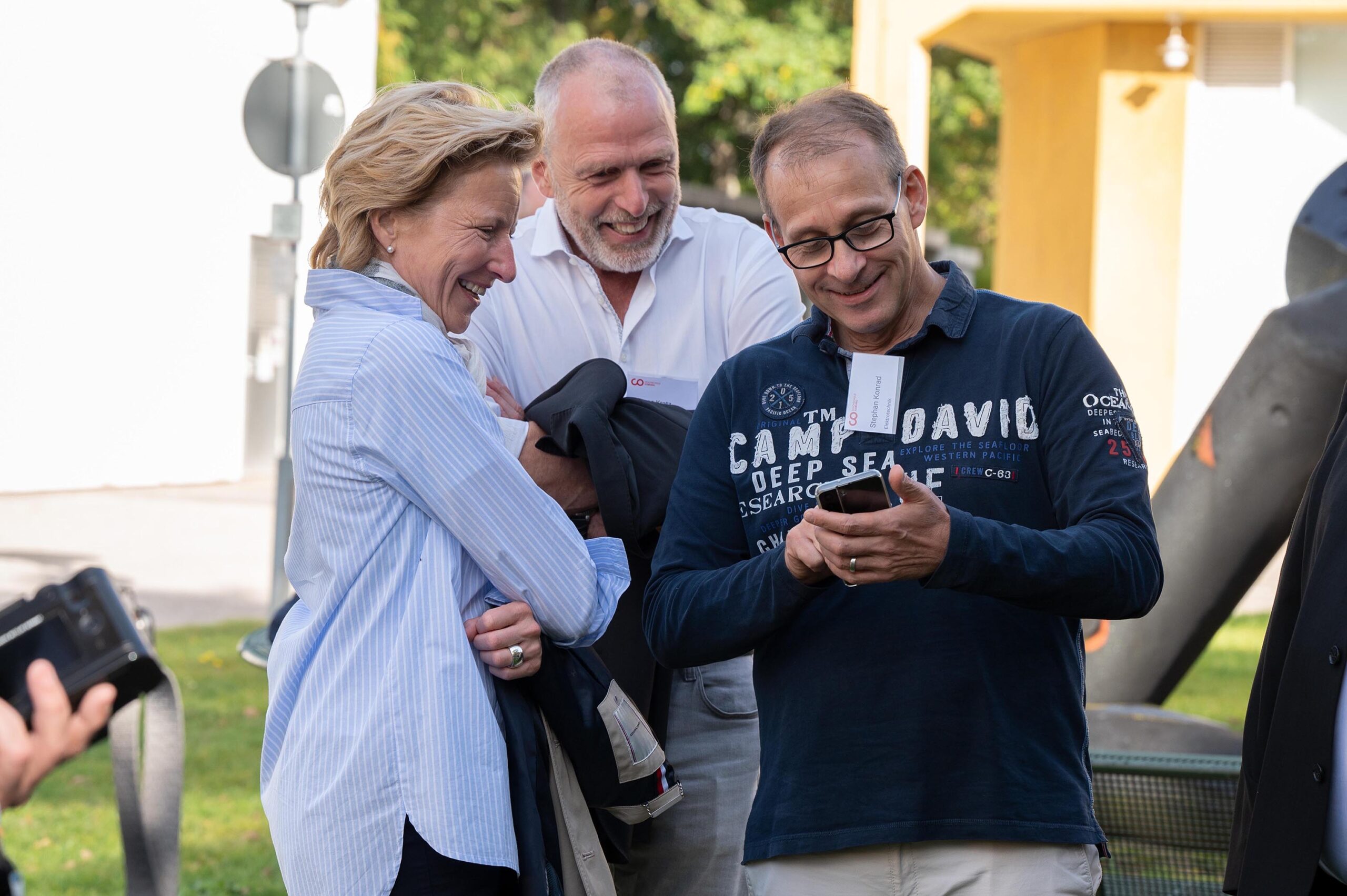 Drei Personen stehen im Freien und lächeln in ein Smartphone, das ein Mann im dunkelblauen Hemd hält. Eine Frau im blauen Hemd hält eine Jacke und hinter ihnen steht ein Mann im weißen Hemd. Im Hintergrund sind Bäume und die gelbe Fassade der Hochschule Coburg zu sehen.