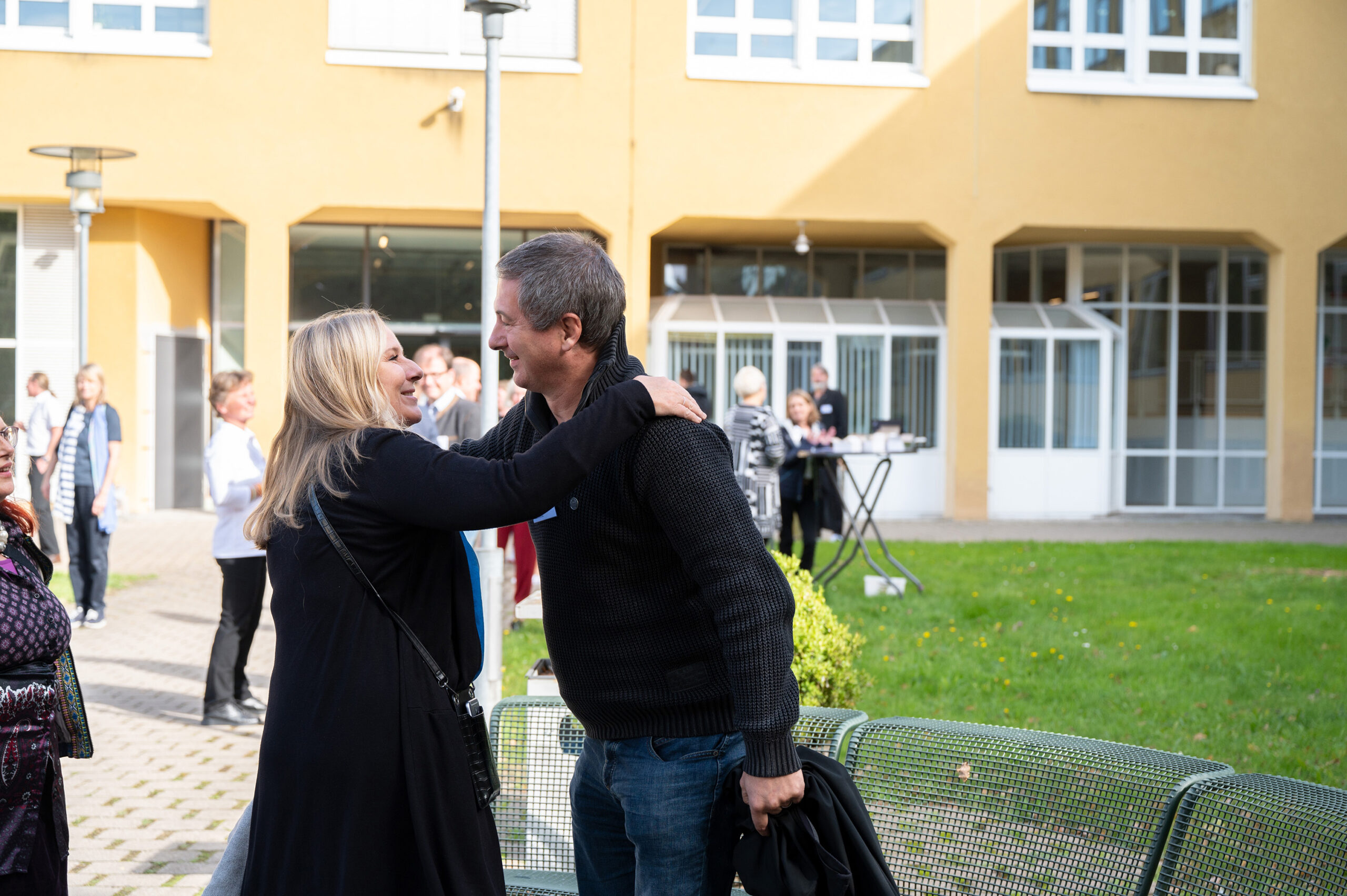 Eine Frau und ein Mann begrüßen sich herzlich vor einem Gebäude der Hochschule Coburg. Sie lächeln und scheinen sich zu umarmen. Im Hintergrund sitzen andere und genießen den sonnigen Tag.