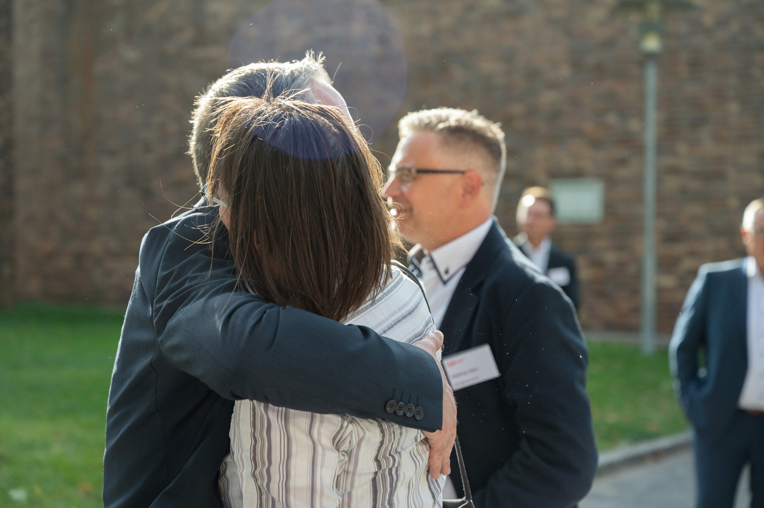 Zwei Menschen umarmen sich herzlich im Freien in der Nähe der Hochschule Coburg, im Hintergrund eine Steinmauer. Eine weitere Person im Anzug steht in der Nähe, leicht unscharf, und blickt in ihre Richtung. Sonnenlicht erzeugt einen subtilen Linsenreflexionseffekt.