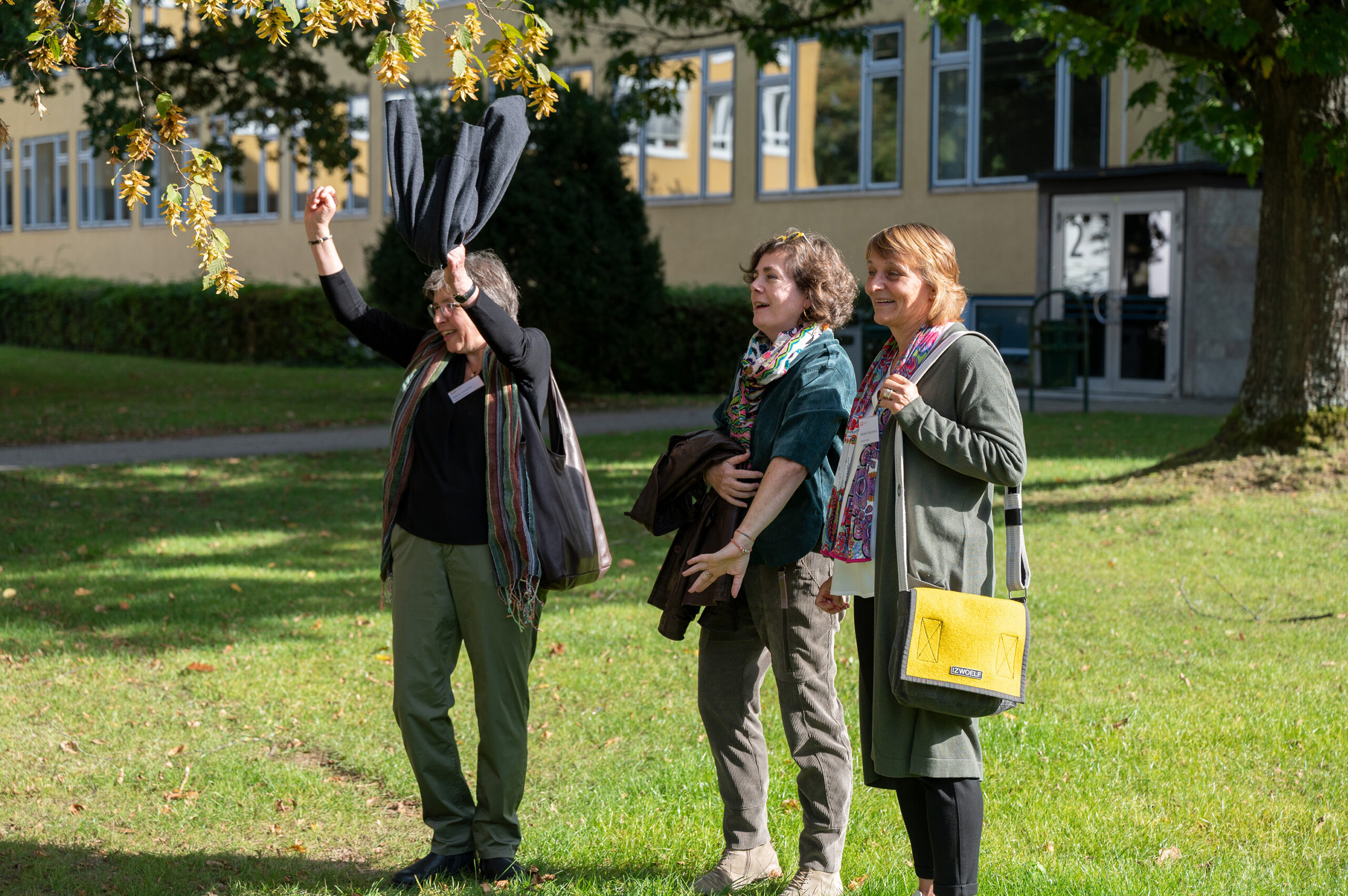 Drei Menschen stehen an einem sonnigen Tag auf der Wiese unter einem Baum in der Nähe der Hochschule Coburg. Eine Person schwenkt verspielt einen Schal in der Luft, während die anderen beiden lächeln und Taschen halten. Als Hintergrund dient ihnen ein Gebäude mit vielen Fenstern.