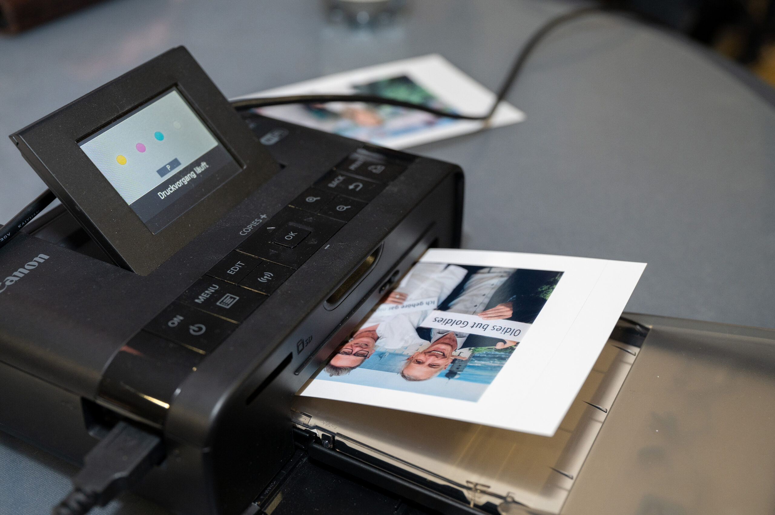 Auf einem grauen Tisch druckt ein kleiner Fotodrucker der Hochschule Coburg ein Bild von zwei Männern. Sein Display zeigt den Tintenstand an, daneben liegt ein verblasstes, buntes Foto.